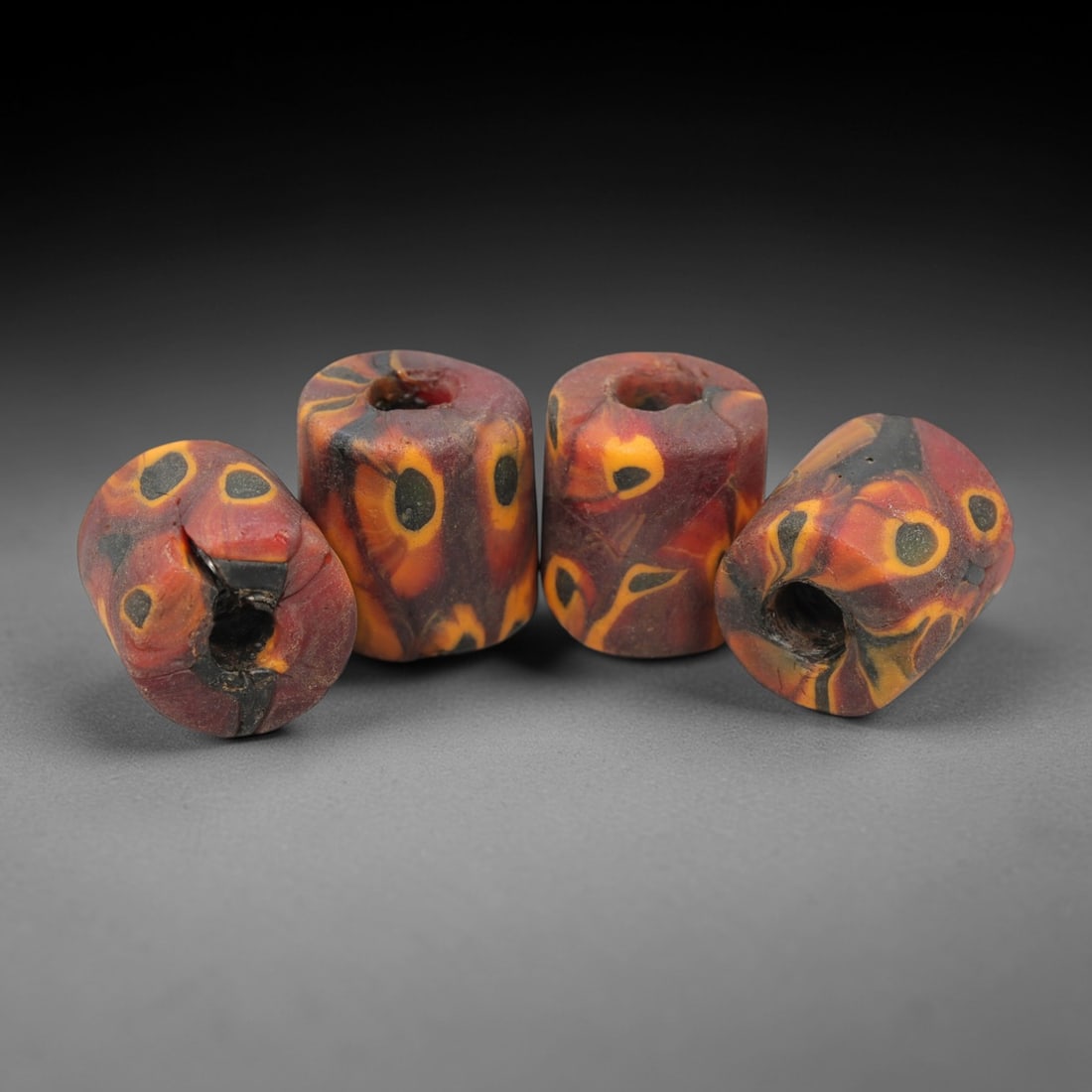 A Set of Four Venetian Millefiori Glass Beads, 19th-20th Century, 78AD: This set of four beads is crafted from opaque red glass, formed as short cylinders with central drilled perforations. The surface is decorated with characteristic millefiori canes, featuring eye-like