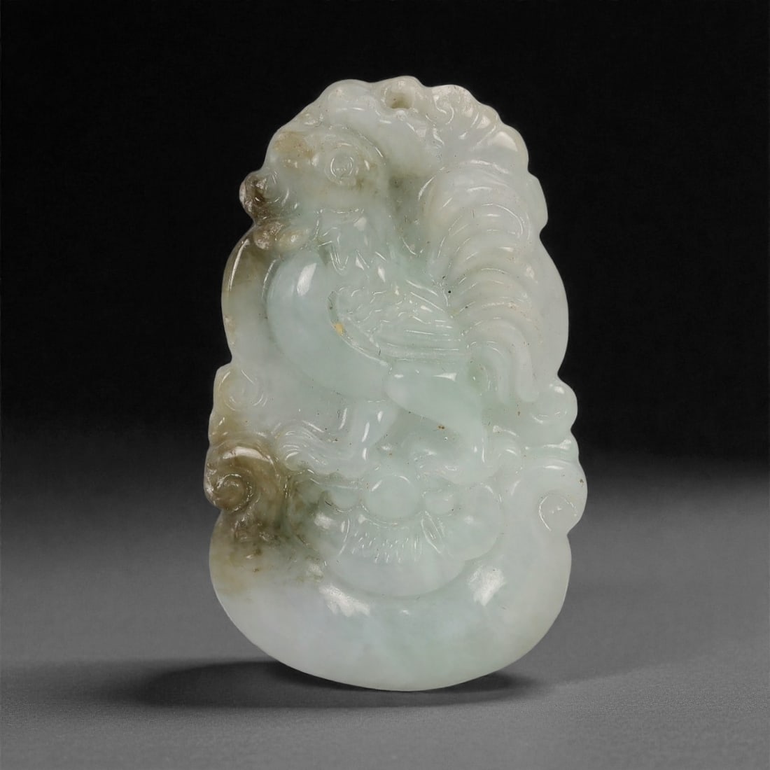 A Chinese Jadeite Jade Chick Amulet Pendant, 20th Century, 45YB: Carved from pale celadon-toned jadeite with areas of creamy translucency and localized brown inclusions, the pendant depicts a stylized chick shown in profile, perched amid softly rendered cloud or fo