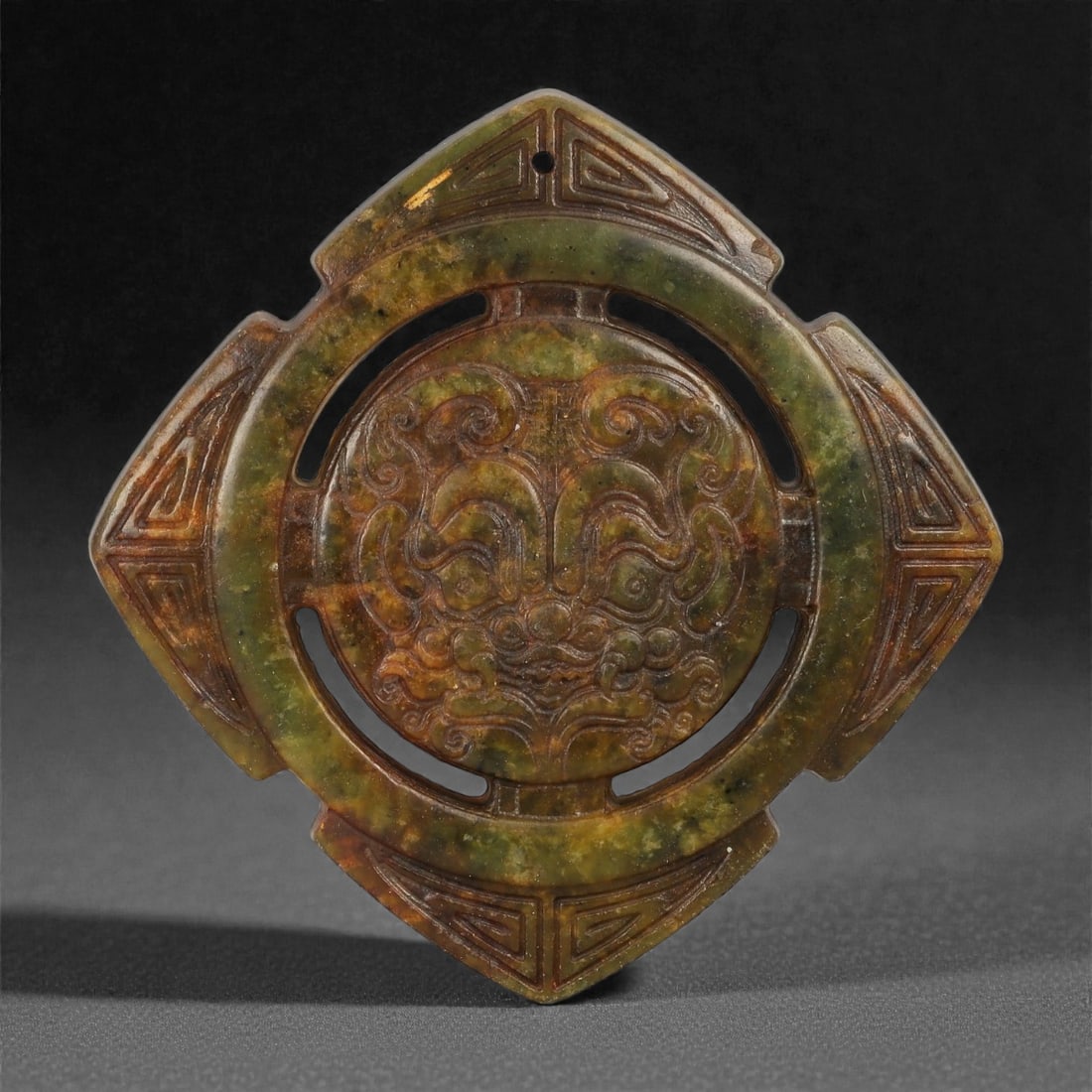 A Chinese Mottled Jade Openwork Disc, 20th Century, 37FD: Carved from mottled green and brown jade with areas of translucency and darker mineral inclusions. The disc is worked in openwork and low to moderate relief, centered with a stylized mask motif render
