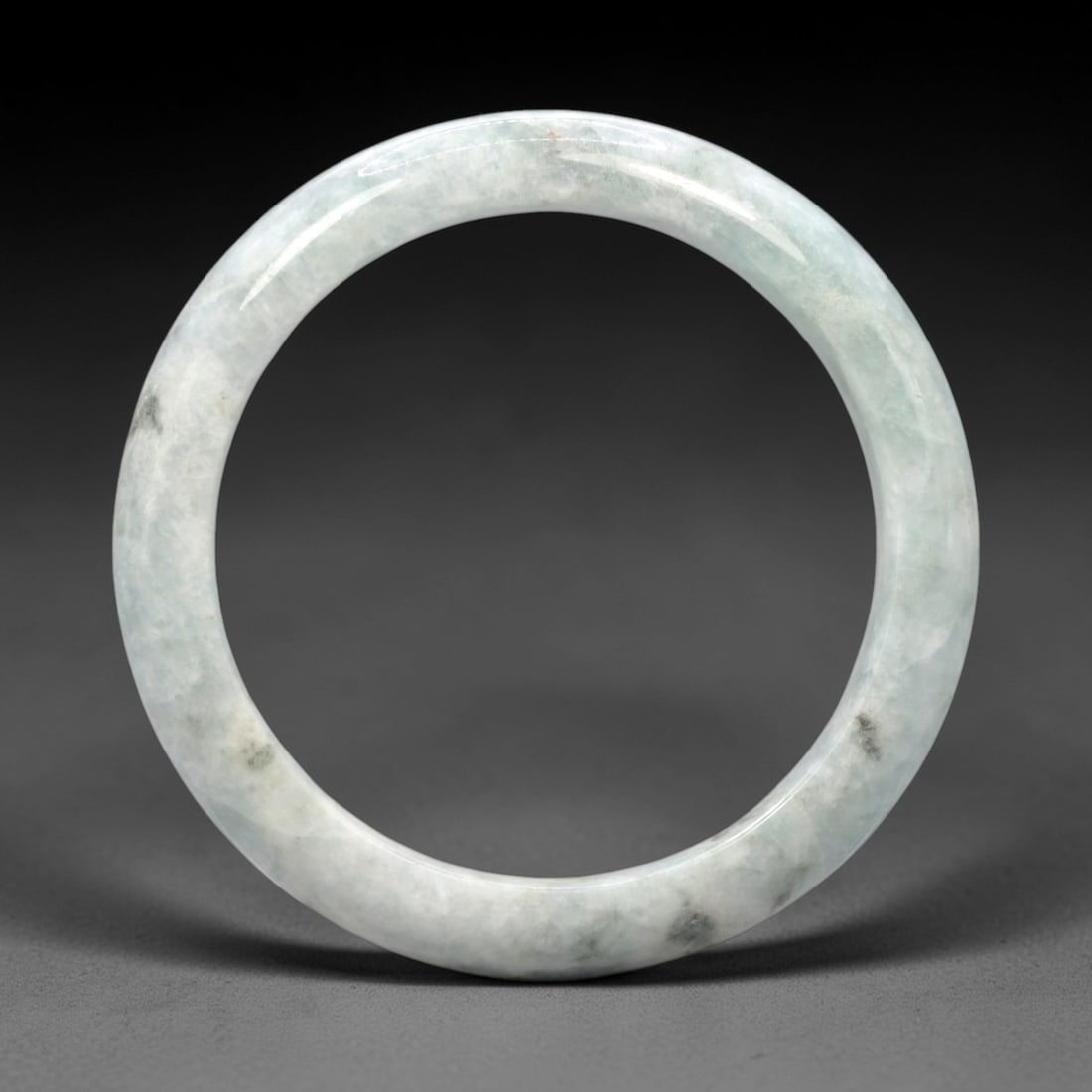 A Chinese Jadeite Jade Bangle Bracelet, 20th Century, 5ZL: Carved from jadeite jade with a pale milky-green base and scattered dark grey inclusions, the bangle is formed as a continuous circular band with a rounded cross-section. The surface is smoothly polis