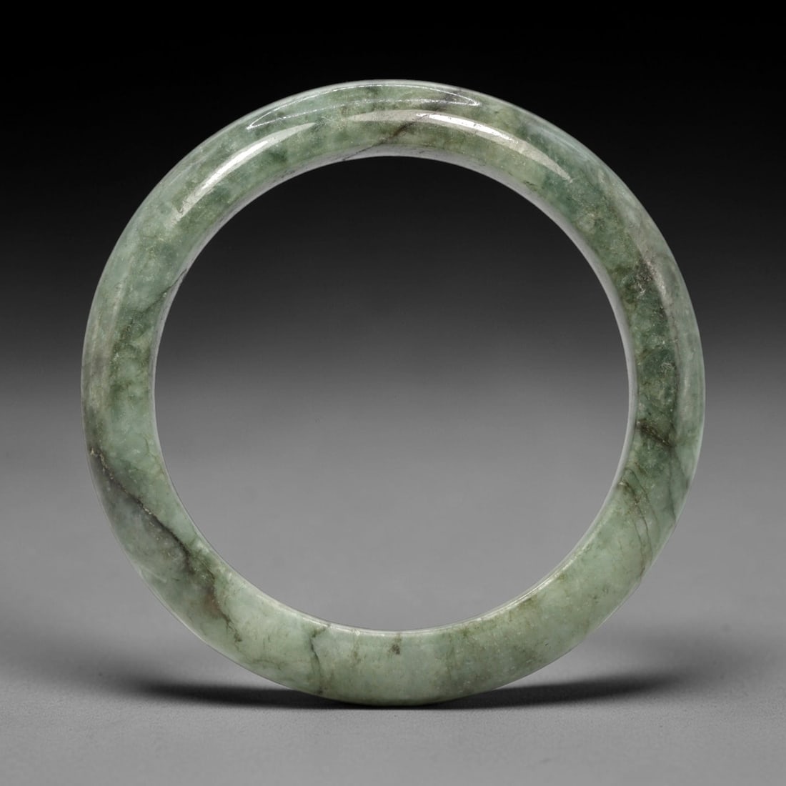 A Burmese Jadeite Jade Bangle, 20th Century, 6FS: A Burmese jadeite jade bangle, with a pale green matrix and prominent dark green mineral inclusions. The bangle is of circular form, with a smooth, polished surface revealing the internal crystalline