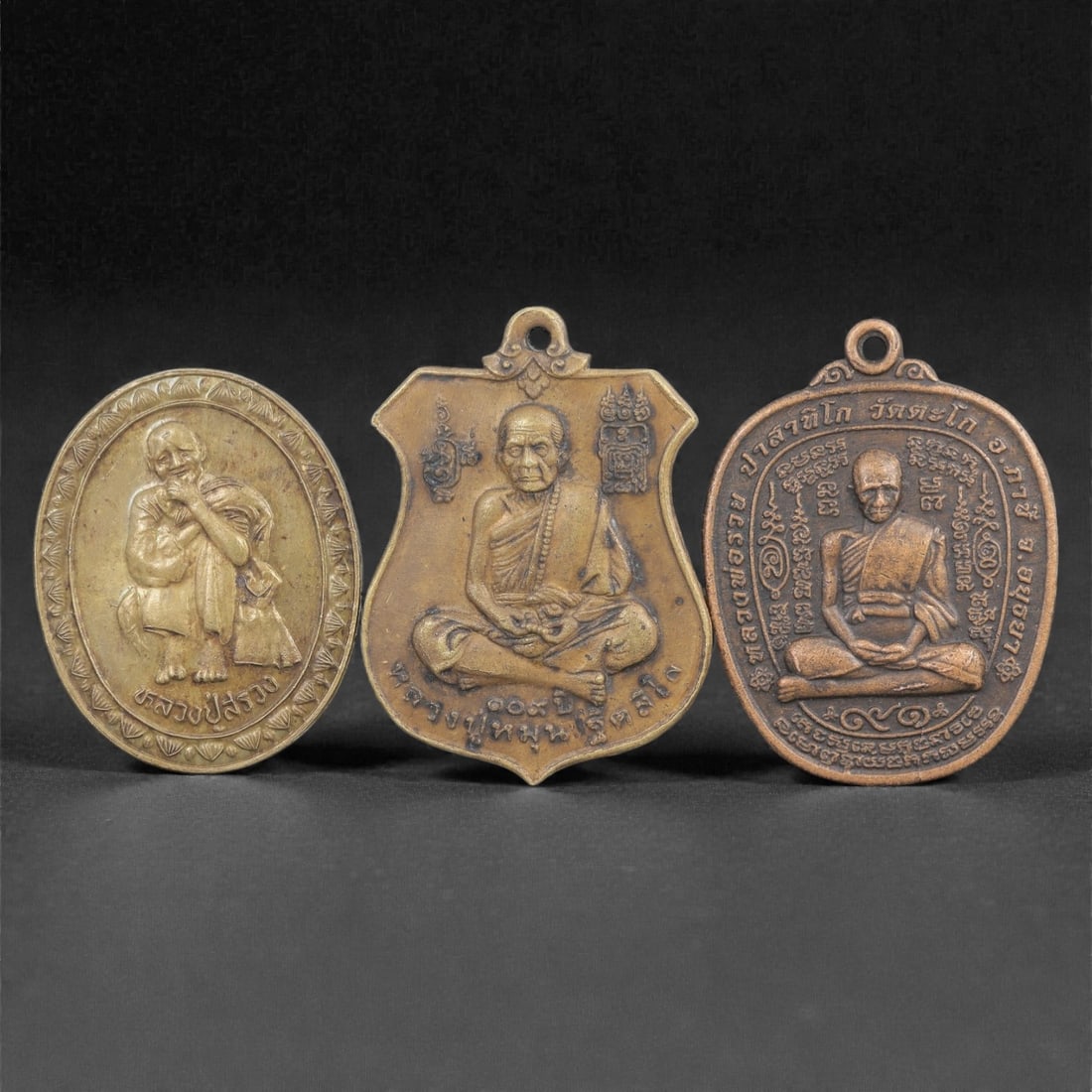 Three Thai Copper Alloy Devotional Amulets, 20th Century, 78FN: Cast from copper alloy with varying warm golden-brown patinas, the group comprises three devotional amulets of distinct forms, each with a suspension loop at the apex for wearable use. The first amule
