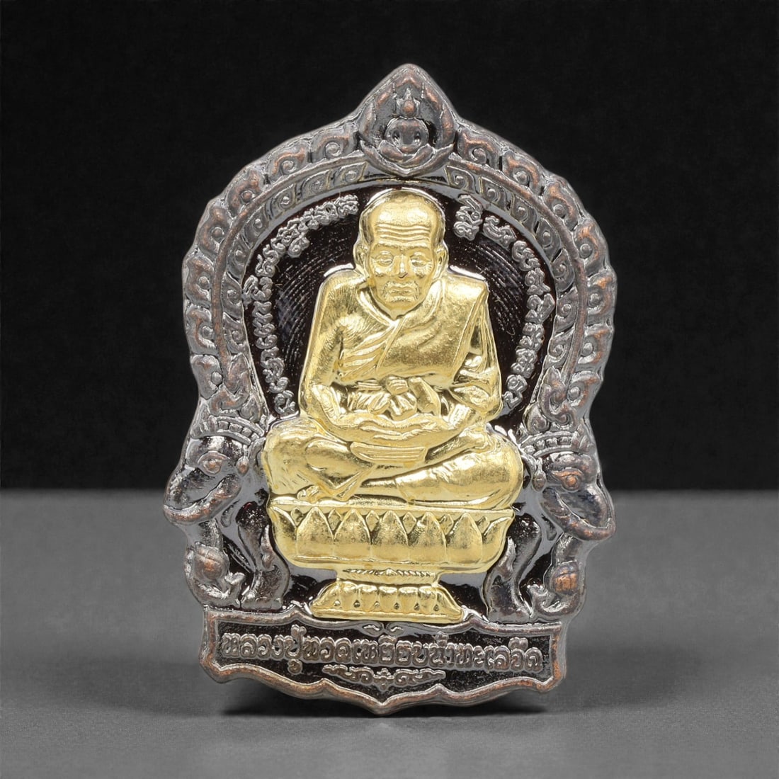 A Thai Bi-Metallic Monk Portrait Amulet, 21st Century, 81FP: Cast with a bi-metallic construction, the amulet has a shield-shaped form with a pointed upper apex. The obverse features a high-relief gilt bust portrait of a seated Buddhist monk, set against a dark