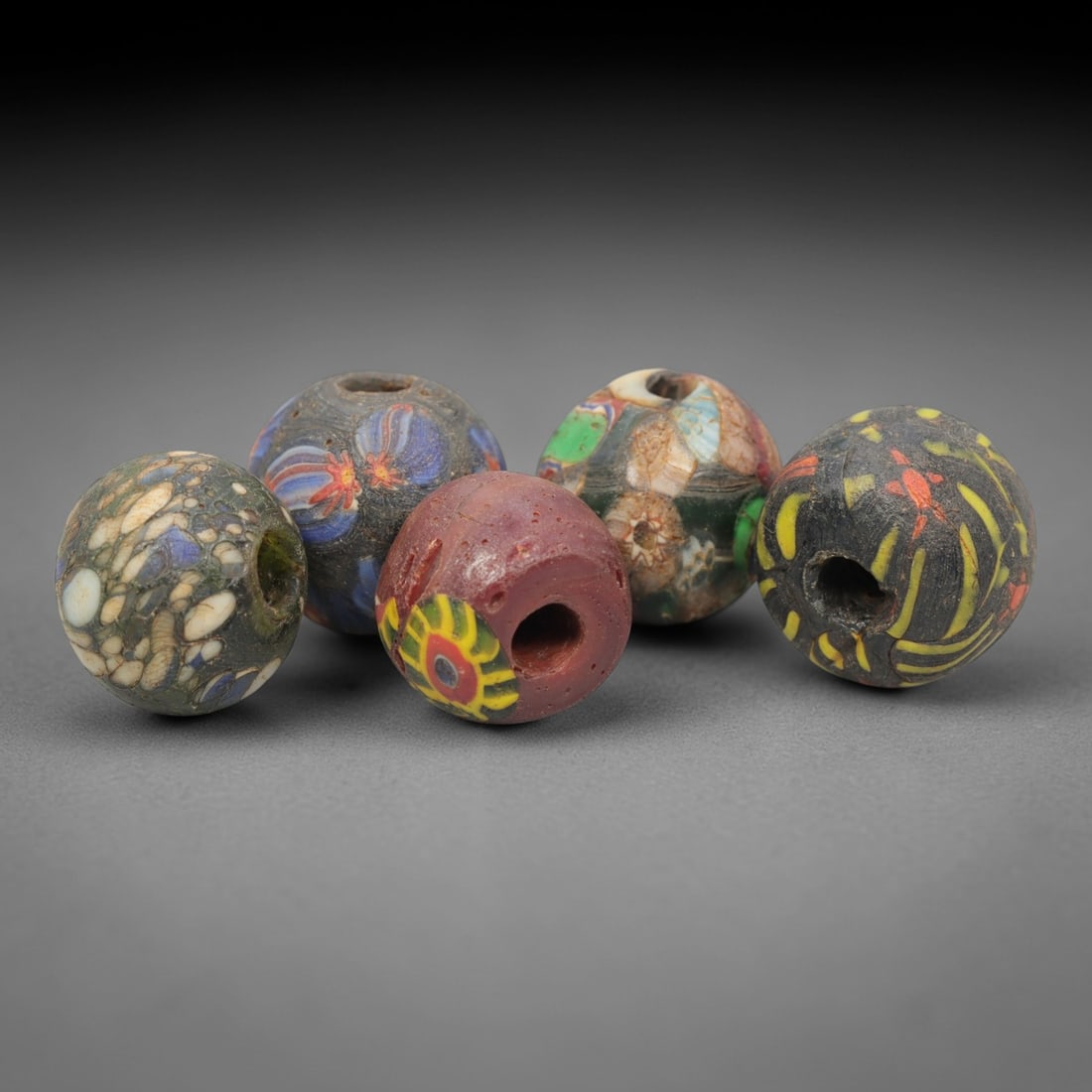 A Group of Venetian Mosaic and Millefiori Glass Beads, 19th-20th Century, 94ZM: Fabricated from opaque and translucent glass, the group consists of five spherical beads with central perforations for suspension. One bead displays a green base with white and blue inclusions; anothe