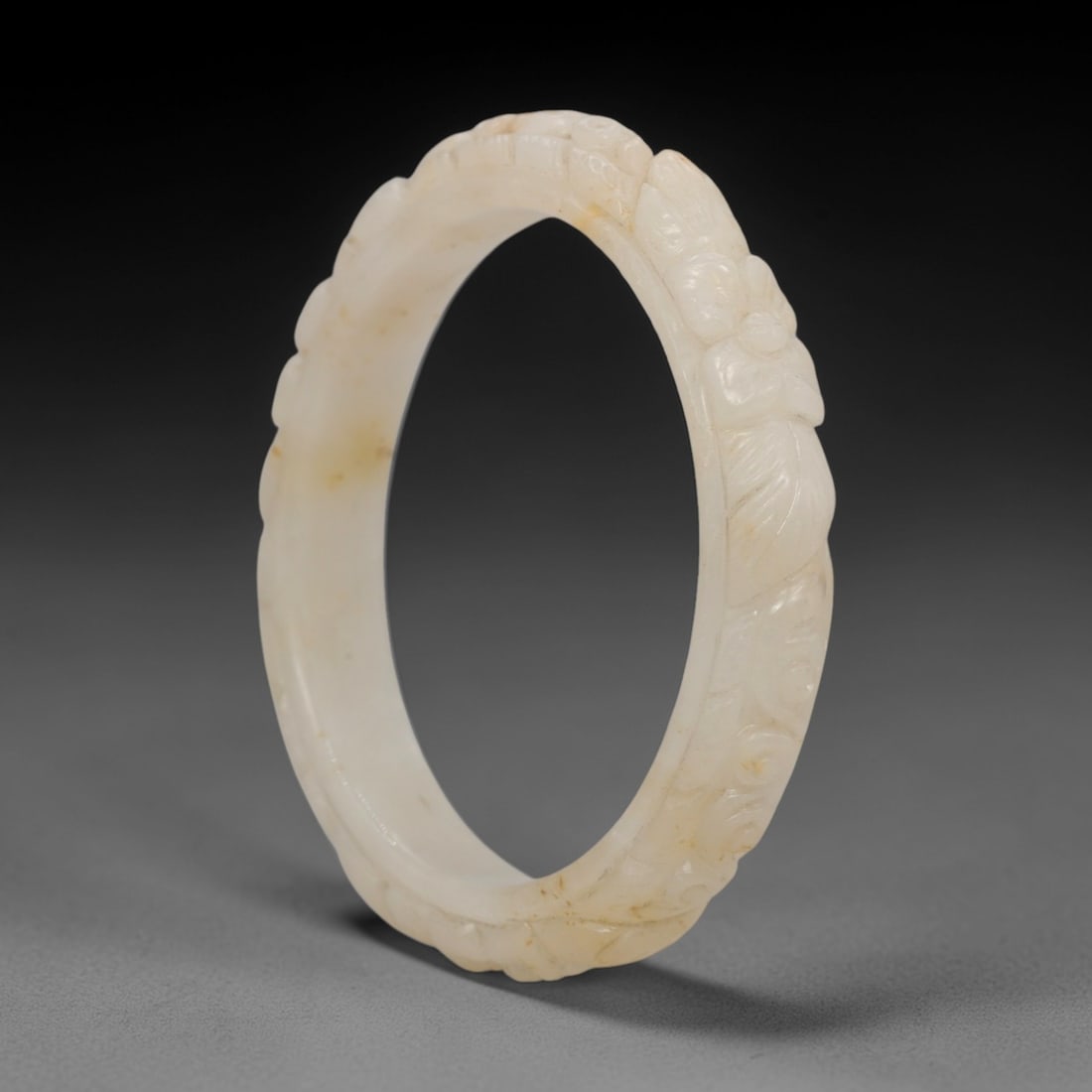 A Chinese Carved Jadeite Jade Bangle Bracelet, 20th Century, 44ZL: A fine jadeite bangle of flattened circular form, masterfully carved in relief with a continuous band of floral blossoms and scrolling foliate motifs. The stone exhibits a creamy white base, accented