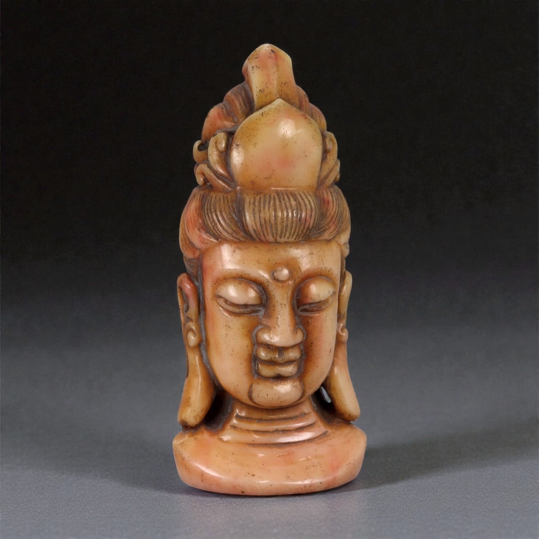 A Chinese Carved Stone Head of a Bodhisattva, 20th Century, 31YB: Carved from a warm-toned stone or hard composite material with mottled beige and light brown coloration, the sculpture represents a bodhisattva head mounted on an integral plinth base. The face is ren