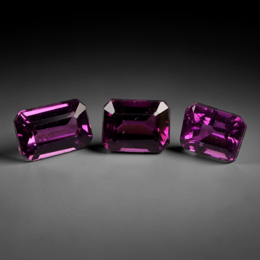 2.24 Carats Deep Raspberry Rhodolite Garnet Parcel - Natural Mozambican Stones, 50XP: This curated parcel of three emerald-cut rhodolite garnets radiates a rich, deep raspberry-purple hue, saturated with vibrant magenta undertones that glow intensely under light. The primary stone meas