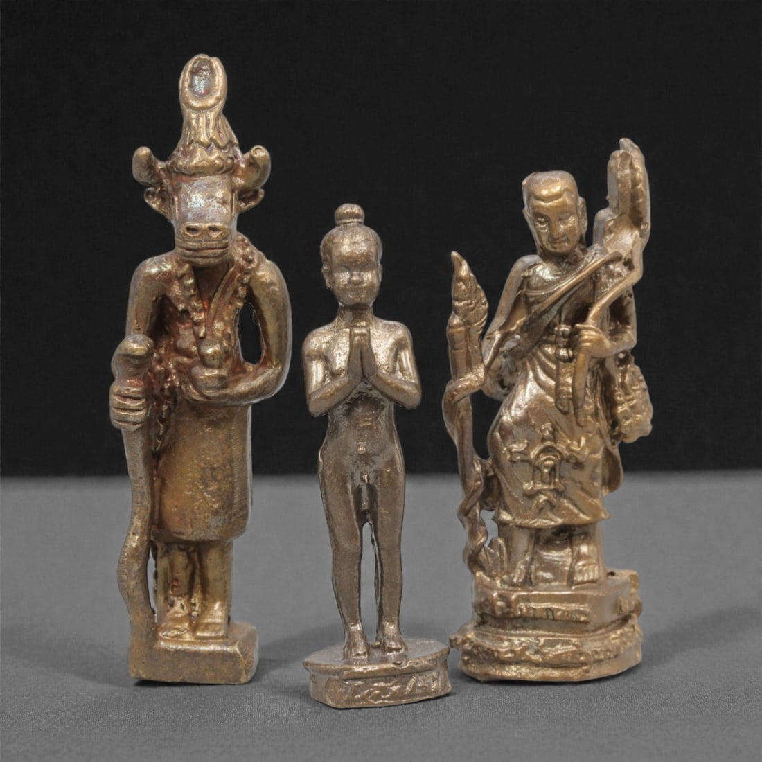 Three Thai Bronze Miniature Figures, 20th Century, 23ZH: Cast from bronze with a warm, aged patina, the set comprises three miniature figures: a bull-headed deity with a headdress and staff, a nude figure in anjali mudra, and a standing monk holding ceremon