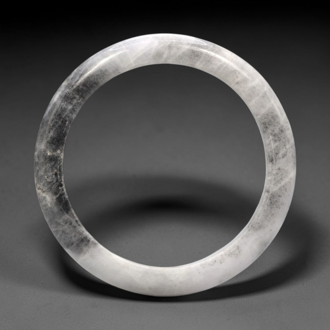 A Chinese Jadeite Jade Bangle Bracelet, 20th Century, 7ZL: Carved from jadeite jade with a translucent pale white base and subtle greyish-black mottling throughout, the bangle is formed as a continuous circular band with a rounded cross-section. The surface i