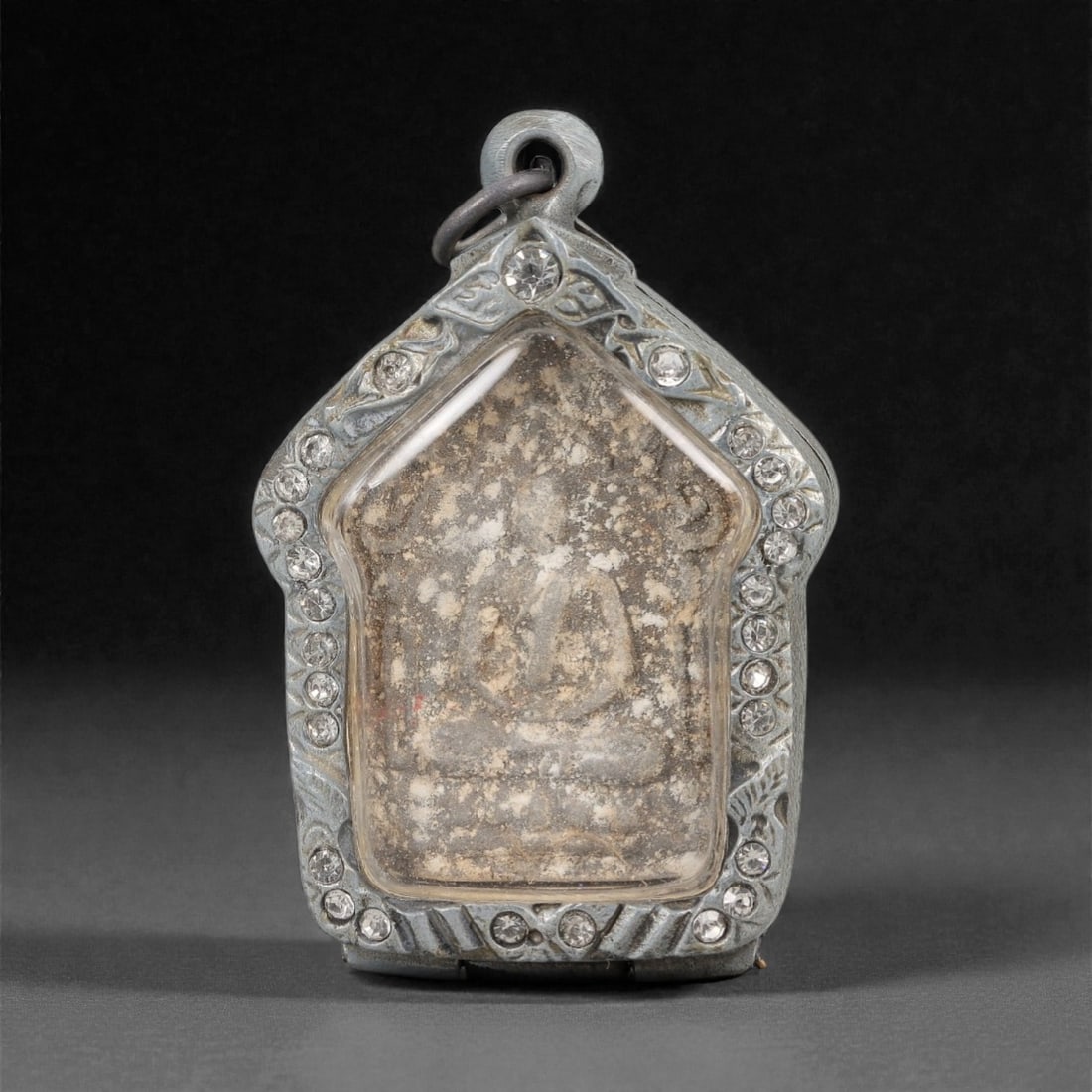 A Thai Buddha Amulet with Metal Frame, 20th Century, 35FP: The amulet is of house-shaped form, featuring a molded seated Buddha figure in meditative pose (dhyana mudra) on the obverse, rendered in a light-colored material against a tan ground, with age-relate