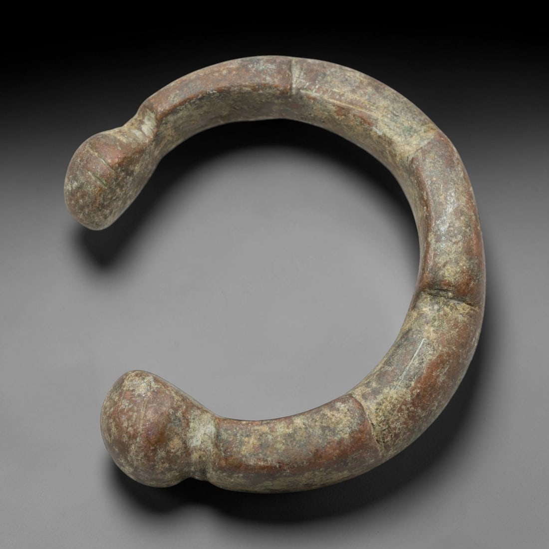 An African Bronze Manila Bracelet, 19th Century, 99FT: Cast from bronze with a thick, rounded section, the bracelet is formed as an open C-shape with prominent, bulbous terminals. The band is segmented by shallow, incised horizontal lines, creating a seri
