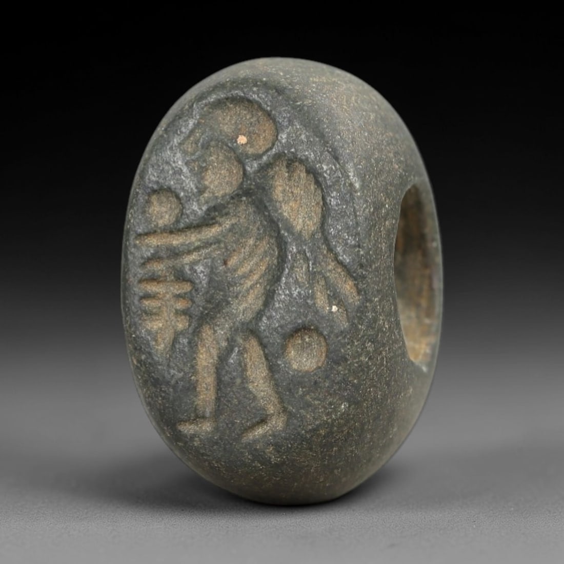 Ancient Stone Hemispherical Stamp Seal - Near Eastern Winged Figure, 29AN: Carved from a dense, dark grey stone, this hemispherical stamp seal features a flat, oval face incised with a standing winged anthropomorphic figure. The figure is depicted with a headdress, a pleated