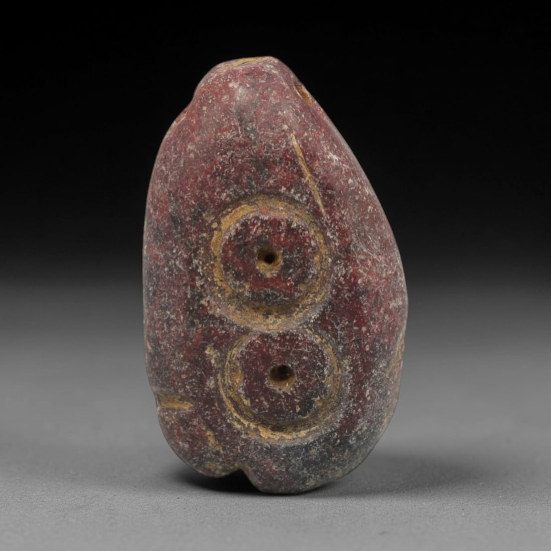 Ancient Stone Amulet - Near Eastern Double Circle Pendant, 13XR: This amulet is carved from a dense, reddish-purple stone, shaped into an elongated teardrop form with a perforated suspension hole at the upper edge, confirming its use as a wearable protective charm.