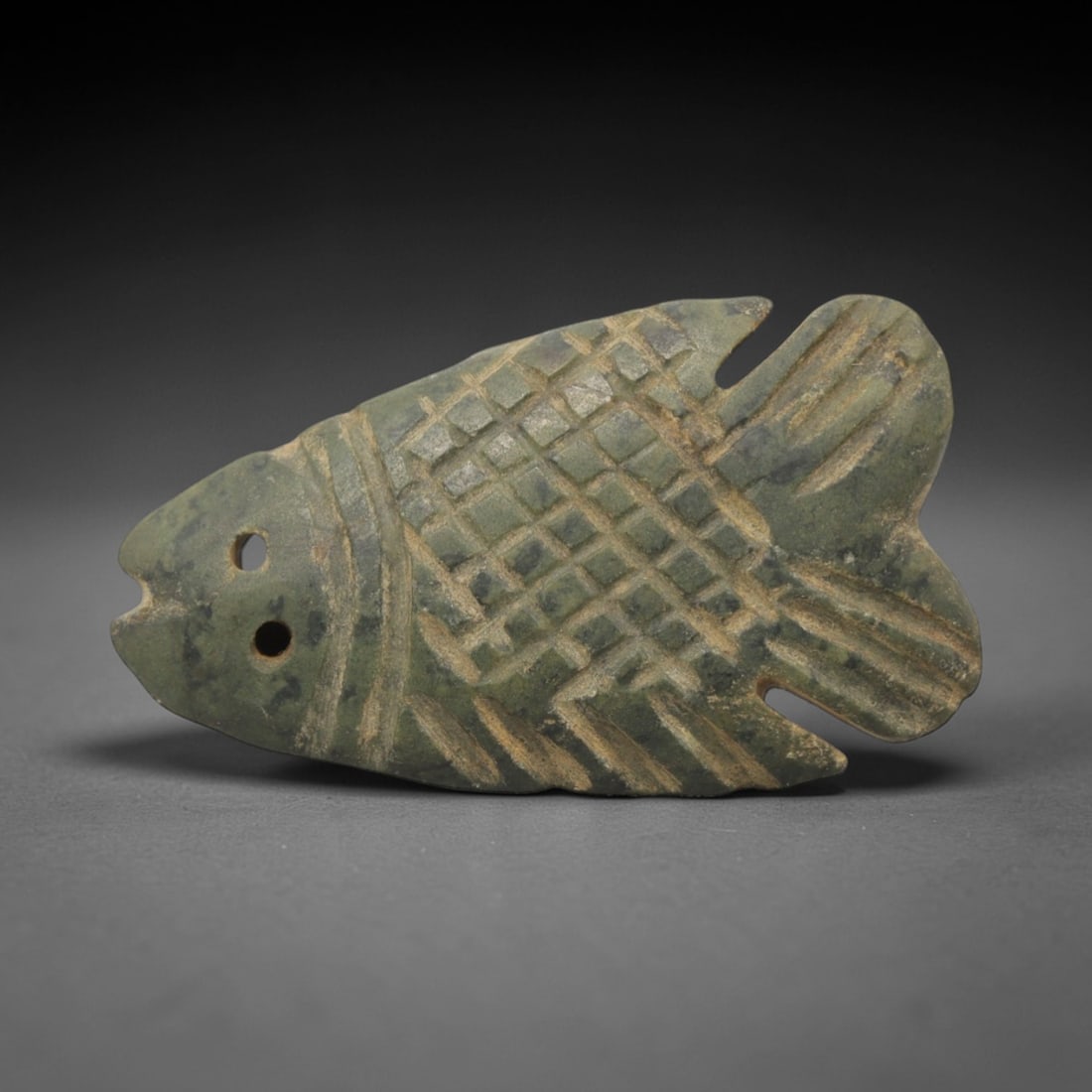 Ancient Stone Fish Amulet - Egyptian Pendant Figurine, 2XR: Carved from a mottled green stone, this amulet depicts a stylized fish with incised grid-patterned scales, linear fin details, and dual perforations at the head for suspension. The form and iconograph