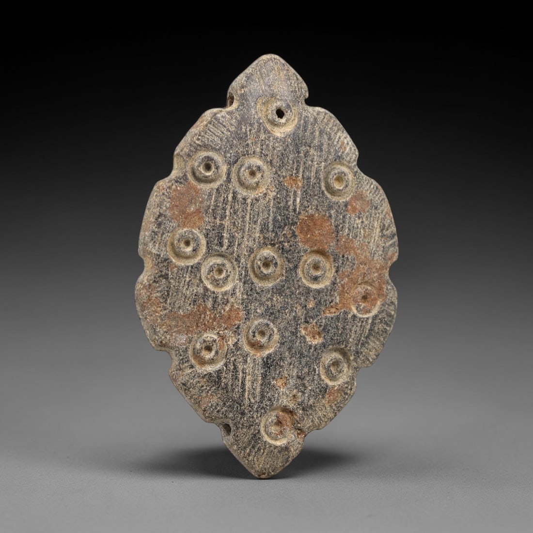 Ancient Stone Amulet - Near Eastern Concentric Circles, 8AL: This amulet is carved from granular stone, shaped into an elongated leaf-like form with serrated edges and suspension holes at the upper and lower extremities, confirming its function as a wearable ta