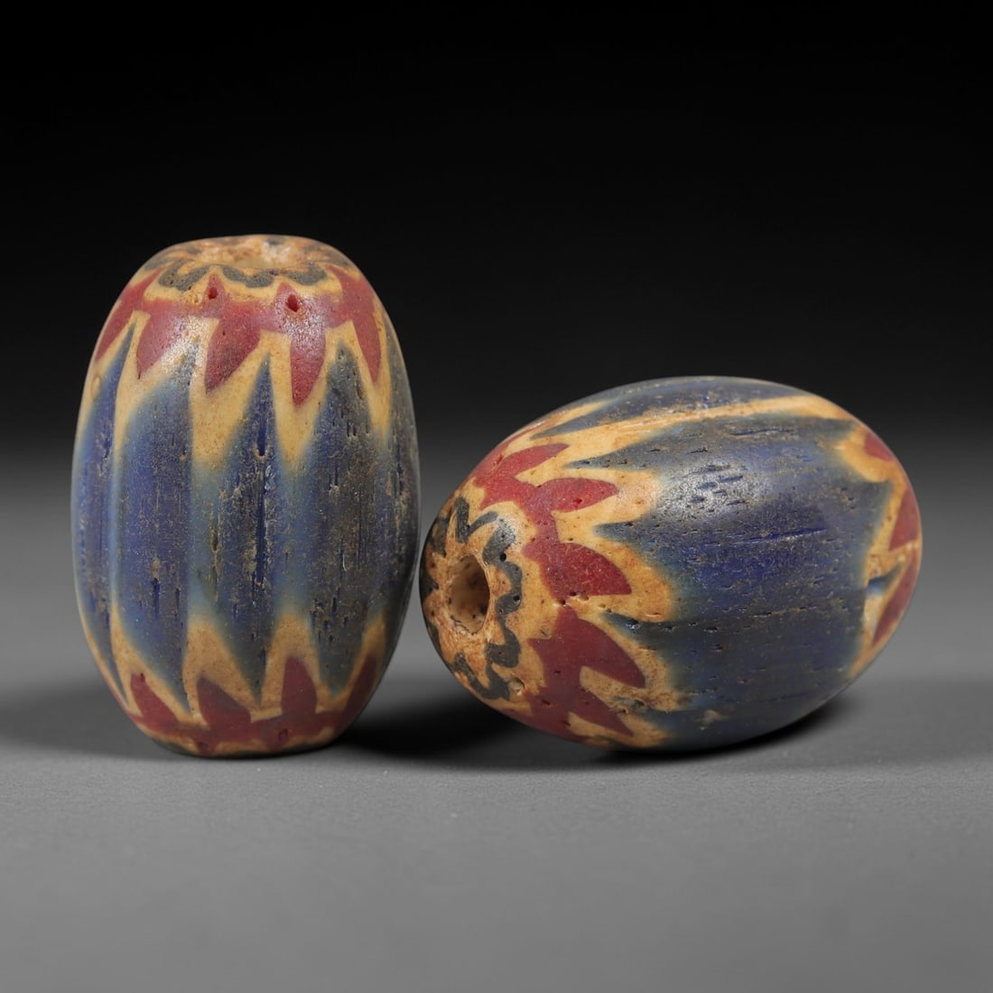 A Pair of Venetian Glass Chevron Beads, 18th-19th Century, 84AC: Crafted from translucent glass with a pale yellow core, each bead is decorated with a chevron pattern of alternating deep blue and vibrant red layers, radiating from the central perforation to the out