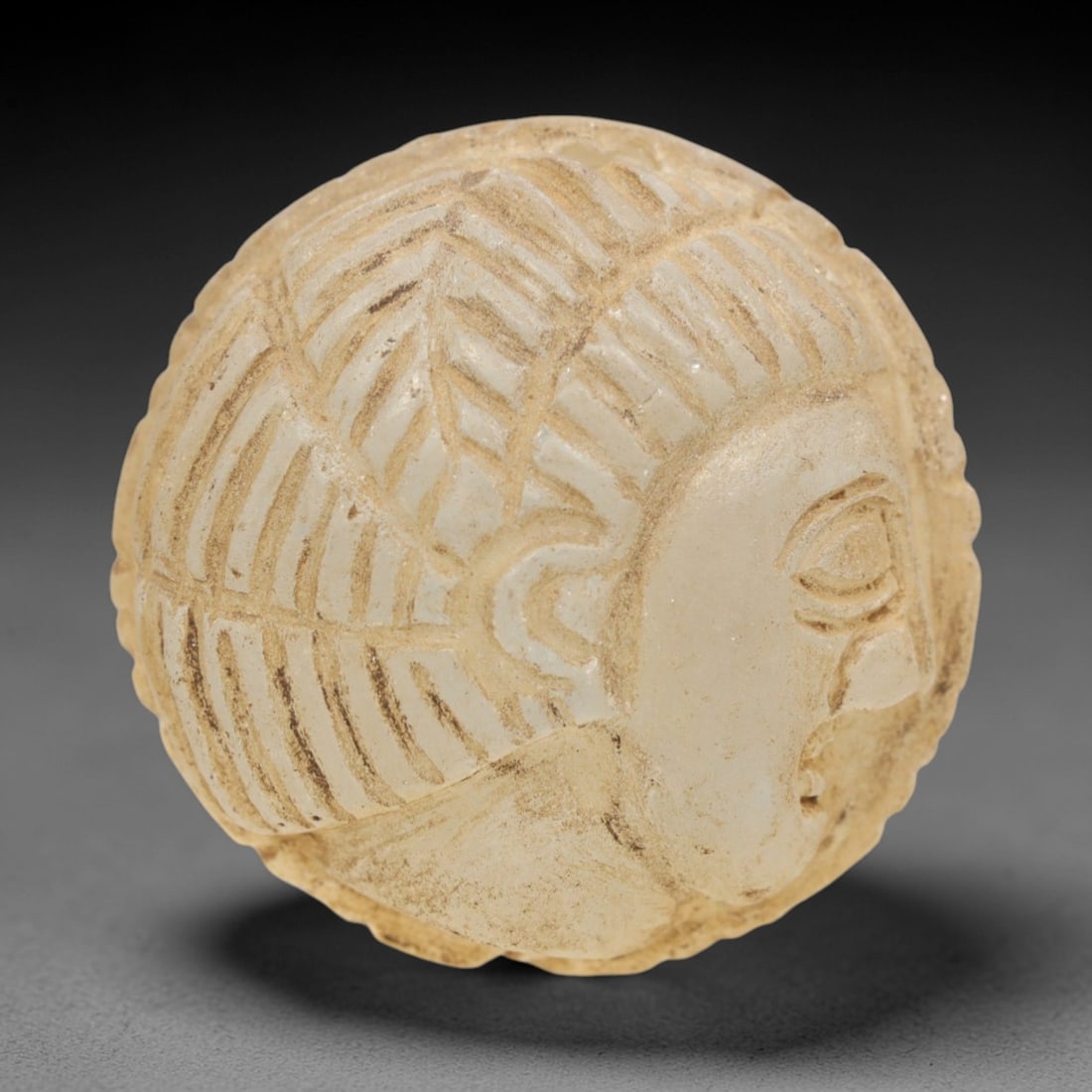 Ancient Glass Medieval Amulet - Near Eastern Bust Figure, 72ZR: Crafted from translucent pale glass with earthy encrustations, this spherical amulet features a raised relief profile bust of a human figure, with stylized linear details defining the geometric hair p