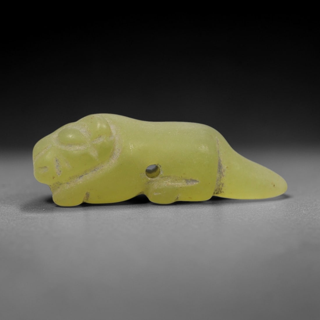 Ancient Serpentine Feline - Near Eastern Amulet Figurine, 74ZW: This amulet is carved from pale yellow-green serpentine, shaped into a stylized recumbent feline, a motif associated with protection and royal authority in ancient Near Eastern iconography. A suspensi