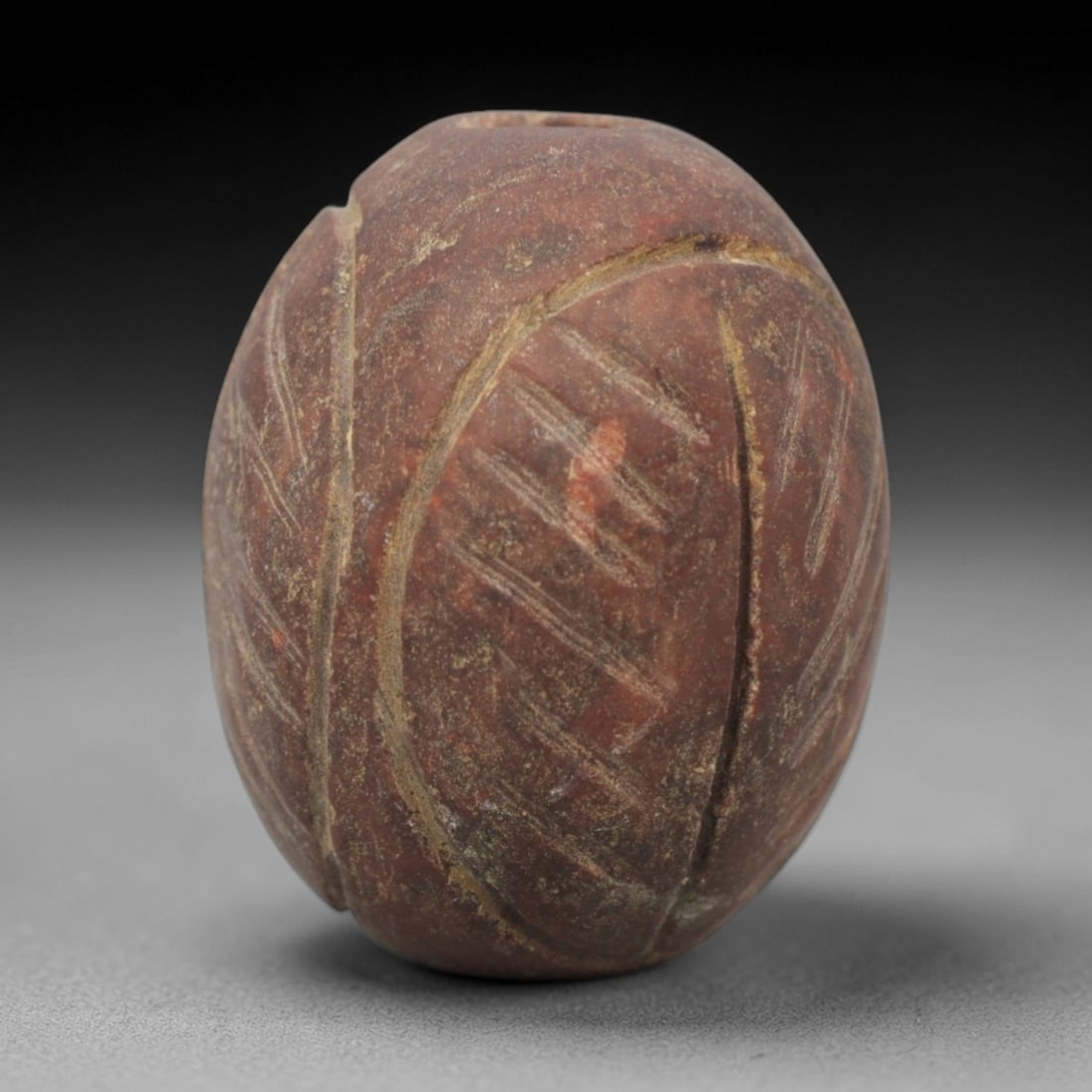 Ancient Red Stone Leaf Amulet - Near Eastern Carved Bead, 11ZV: This amulet is carved from a mottled red stone, formed as an oval bead with incised leaf motifs across its surface. Curved and linear incisions define the leaf veins and outlines, creating a textured