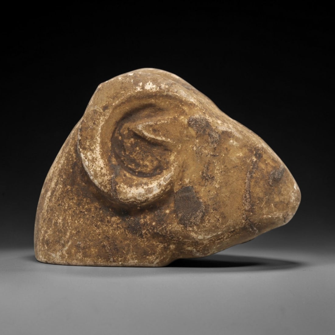 Ancient Stone Zoomorphic Ram Head - Near Eastern Amulet, 67ZV: This artifact is a carved stone zoomorphic ram head amulet, crafted from a granular, earth-toned stone consistent with ancient Near Eastern lithic traditions. The relief carving depicts a stylized ram