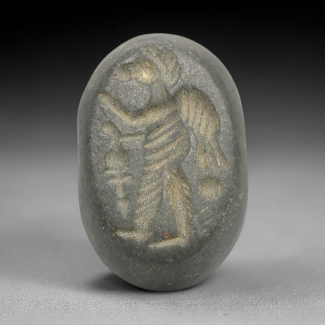 Ancient Stone Hemispherical Stamp Seal - Near Eastern Winged Anthropomorphic Figure, 65ZS: Crafted from dense, dark green stone, this hemispherical stamp seal bears incised carving of a winged anthropomorphic figure, with a bird-like head, segmented torso, and outstretched wings, alongside