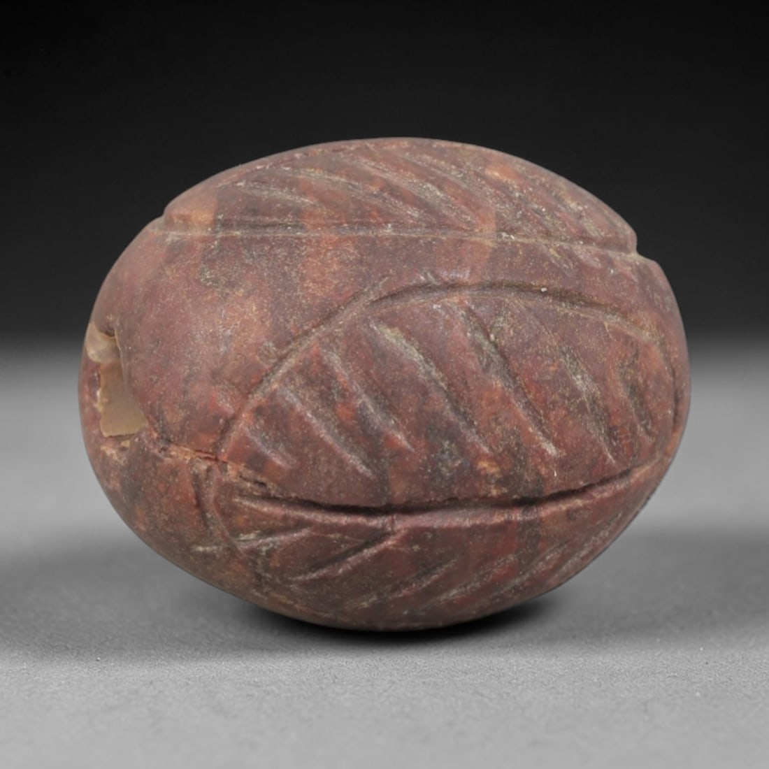 Ancient Stone Bead - Near Eastern Ritual Amulet, 84AP: This artifact is carved from variegated red stone with a weathered, earthy patina and subtle mineral veining, exhibiting incised leaf-like linear detailing consistent with ancient Near Eastern stonewo