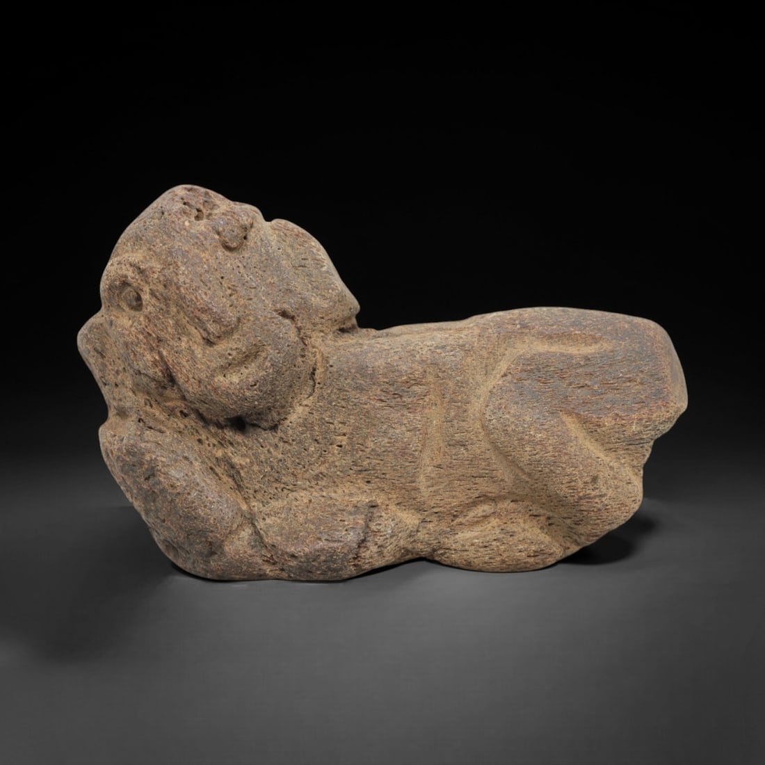 Ancient Stone Recumbent Feline - Mesopotamian Figurine, 27FX: Carved from a coarse, porous stone, this recumbent feline figure exhibits simplified, robust forms with incised lines defining the limbs, facial contours, and ears. The compact, reclined posture and s