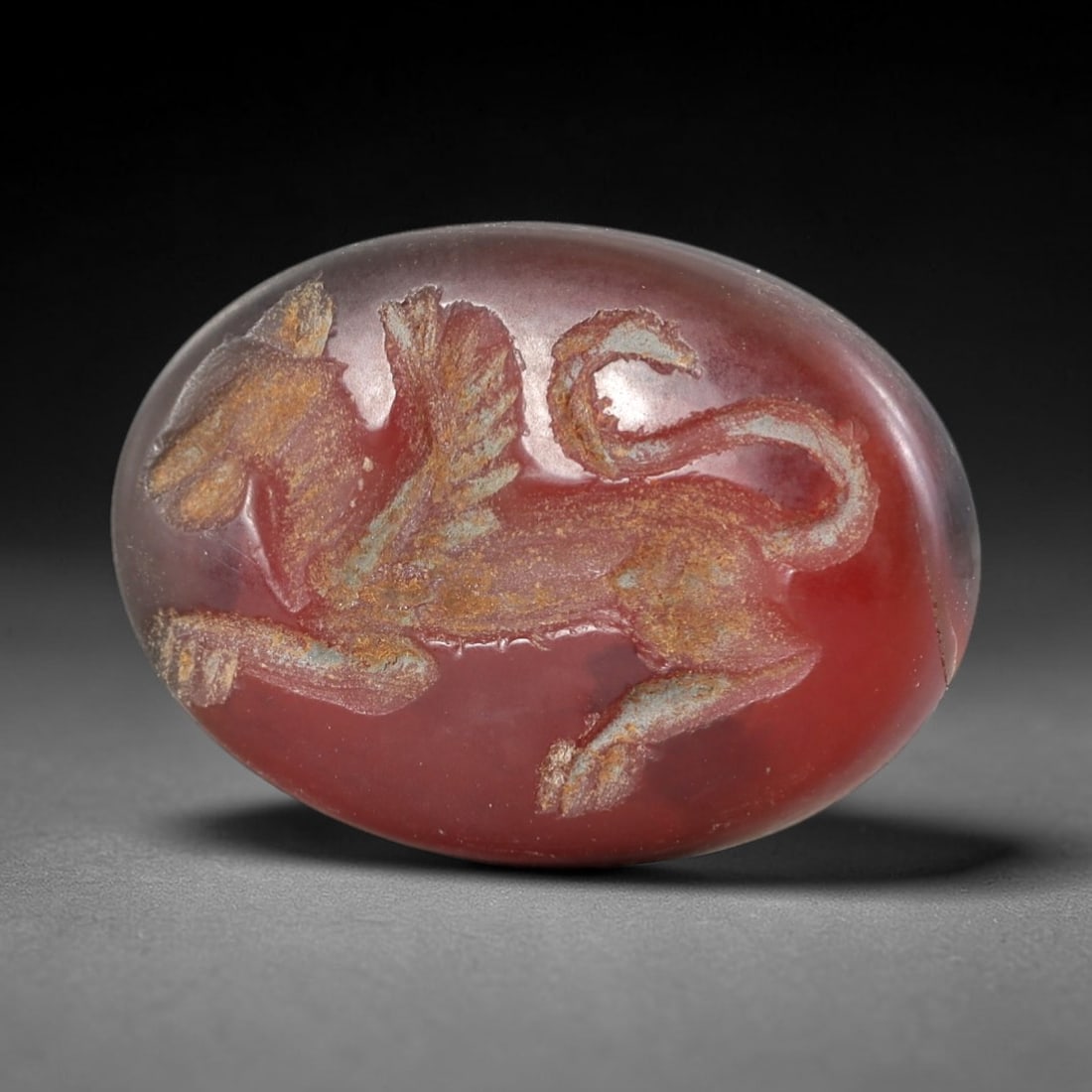 Ancient Agate Griffin Intaglio - Near Eastern Amulet, 78AR: This oval intaglio is carved from deep red agate, featuring a high-relief depiction of a griffin-a mythical hybrid with avian head, feathered wings, and quadrupedal body. The creature is rendered in s