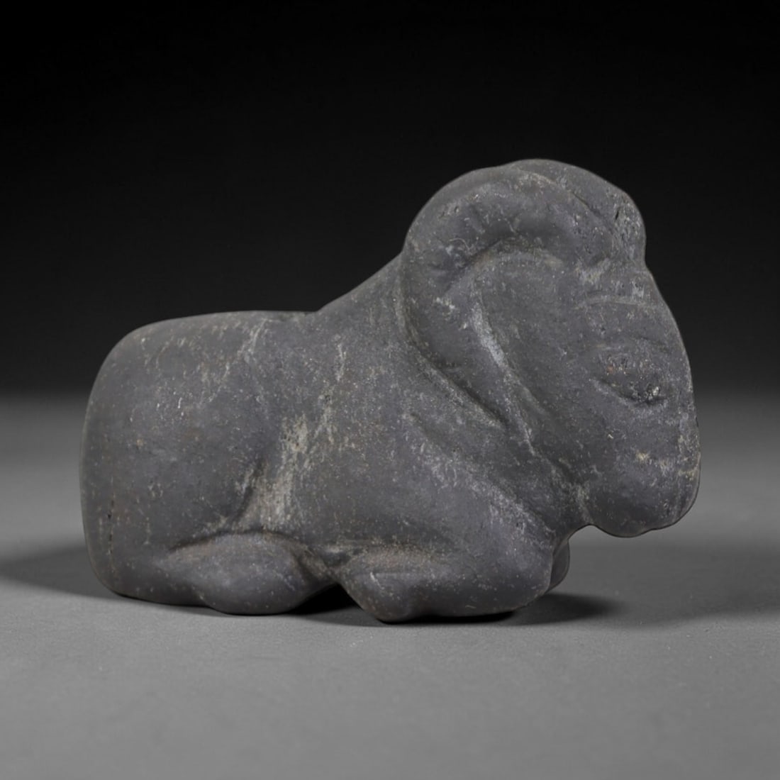 Ancient Stone Ram - Near Eastern Amulet Figurine, 72AP: This artifact is carved from dark gray stone with a weathered, matte patina and subtle surface wear indicative of ancient production. The form depicts a recumbent ram with simplified, stylized feature