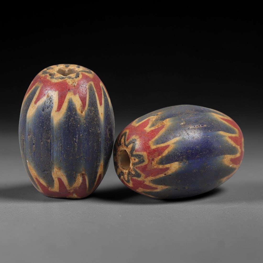 A Pair of Venetian Glass Chevron Beads, 18th-19th Century, 83AC: Crafted from translucent glass with a pale yellow core, each bead is decorated with a chevron pattern of alternating deep blue and vibrant red layers, radiating from the central perforation to the out