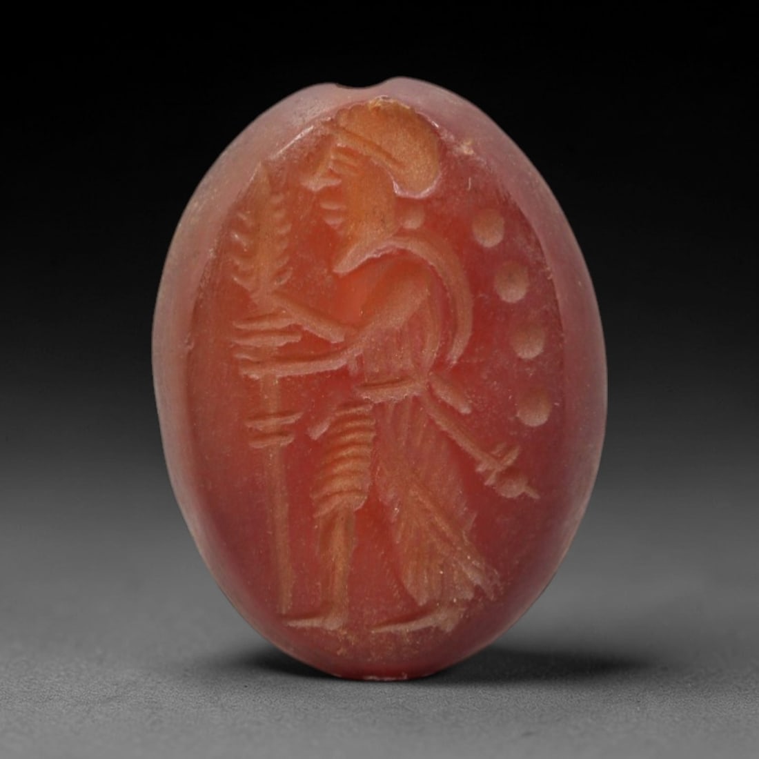 Ancient Carnelian Intaglio - Warrior Figure Seal, 33XV: Crafted from polished carnelian, this oval intaglio seal depicts a standing warrior figure in profile, adorned with a crested helmet and fringed garment. The figure holds a tall, leaf-tipped staff in