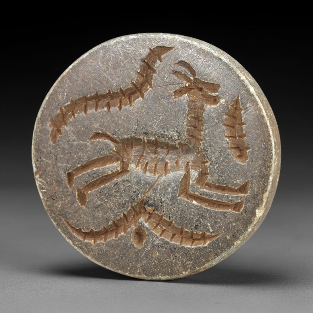 Ancient Stone Stamp Seal - Near Eastern Zoomorphic Impression, 28AM: This circular stamp seal is carved from a dense, weathered stone with a muted, earthy patina indicative of long-term burial exposure. The incised surface features a composite zoomorphic scene: a styli