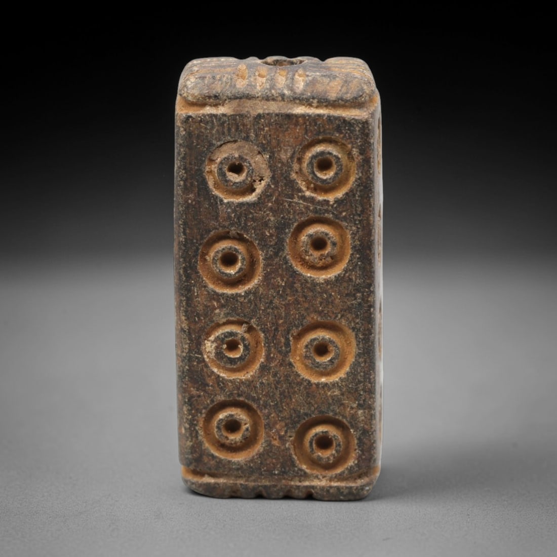Ancient Steatite Amulet Bead - Near Eastern Concentric Circles, 42XV: Carved from dense steatite, this ancient amulet bead features a rectangular prism form with a vertical suspension hole at the top, designed for personal wear. Its face is decorated with a symmetrical