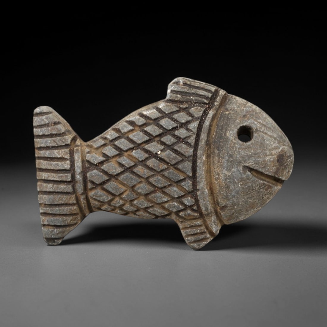 Ancient Stone Fish Amulet - Near Eastern Aquatic Figure, 34AM: This fish amulet is carved from dark speckled stone, depicting a stylized fish form with intricate incised geometric detailing defining the scales, fins, and facial features. The piece reflects the ic