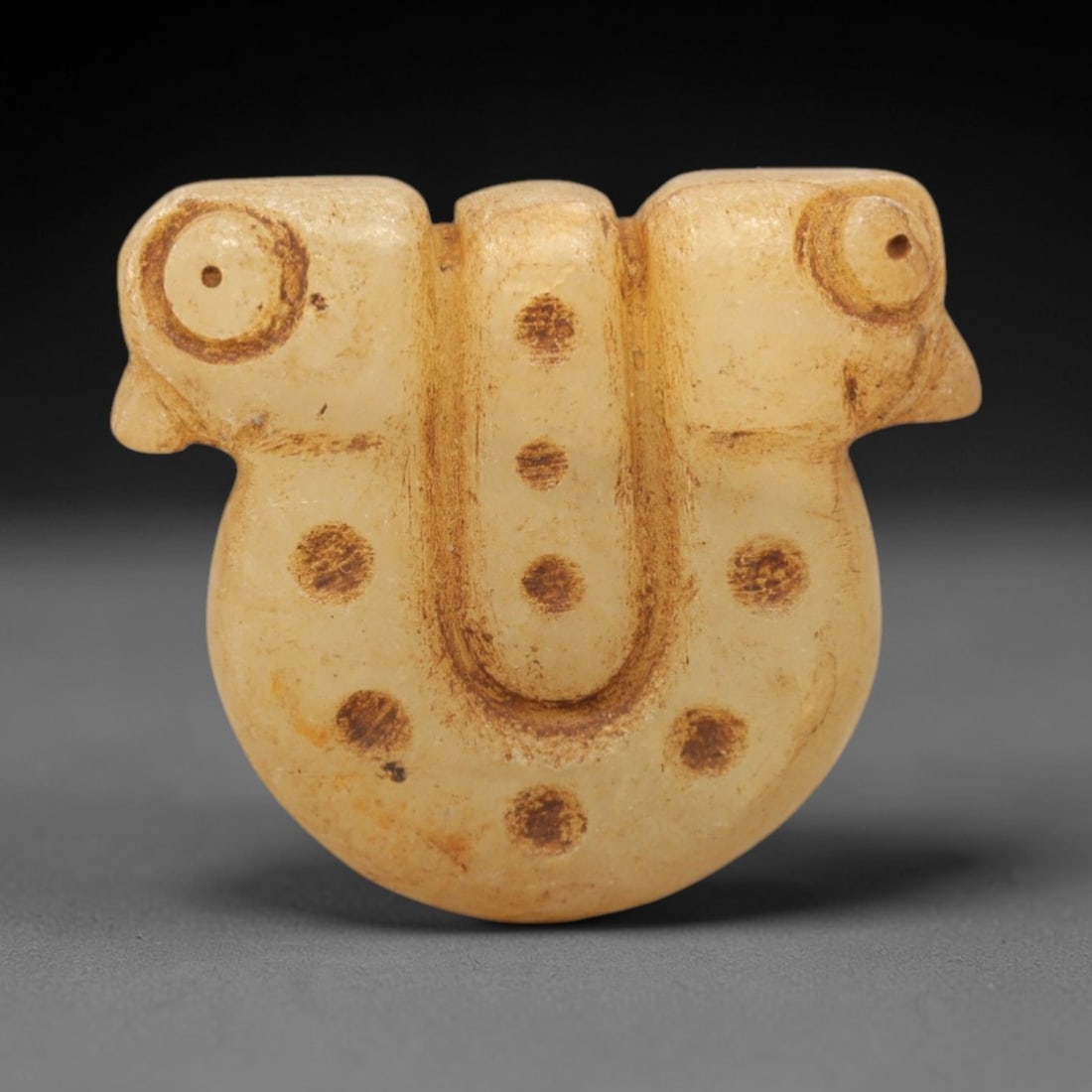 Ancient Chalcedony Double Bird Amulet - Near Eastern Figurine, 28XV: Carved from pale chalcedony, this amulet features a symmetrical double-bird form with two stylized bird heads flanking a central U-shaped ridge, decorated with incised circular dot motifs across the s