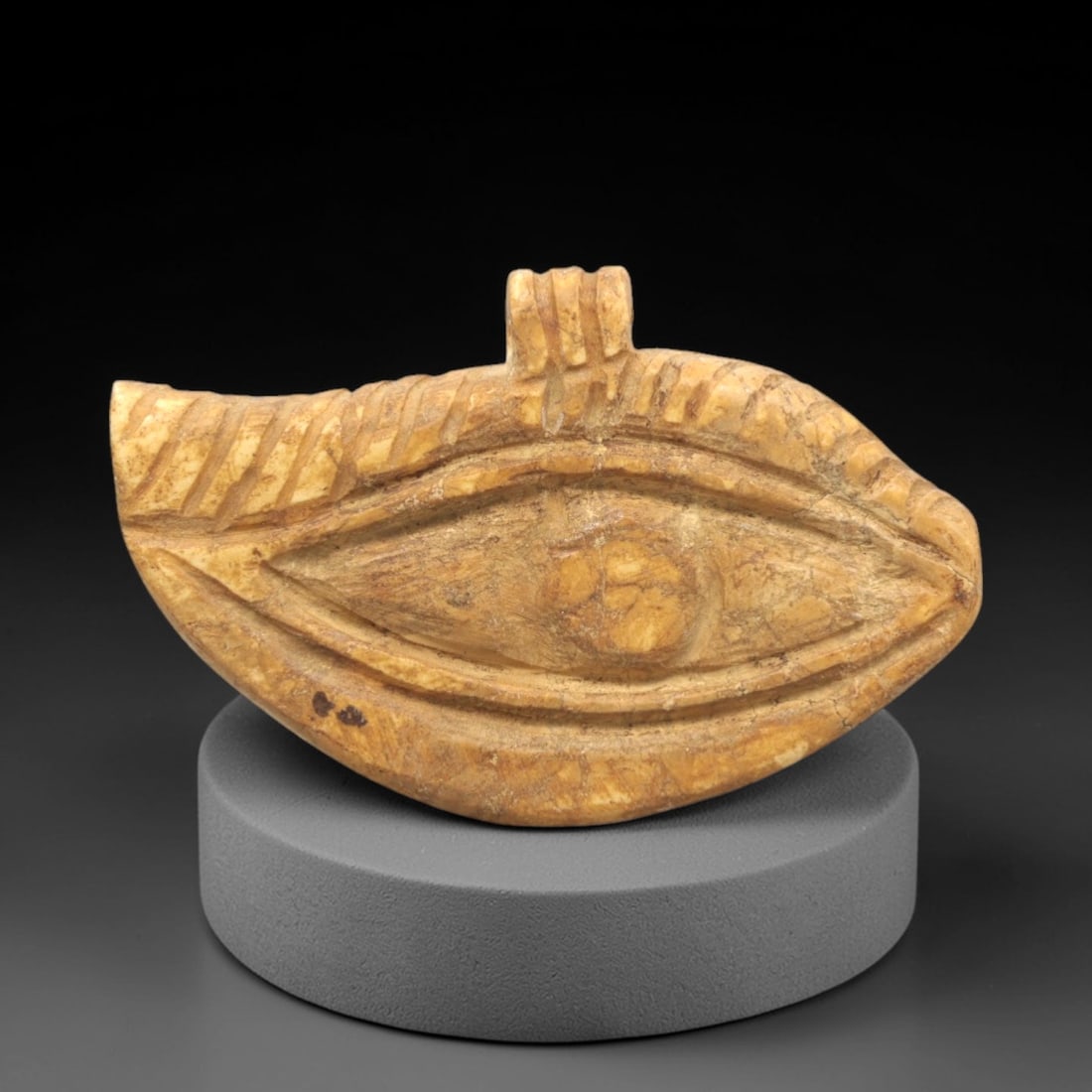 Ancient Stone Wedjat Eye - Egyptian Amulet Pendant, 94ZP: Carved from warm tan stone, this Wedjat Eye amulet features a stylized eye form with incised eyelid and brow lines, a raised central pupil, and a ribbed decorative border along the upper edge. A small