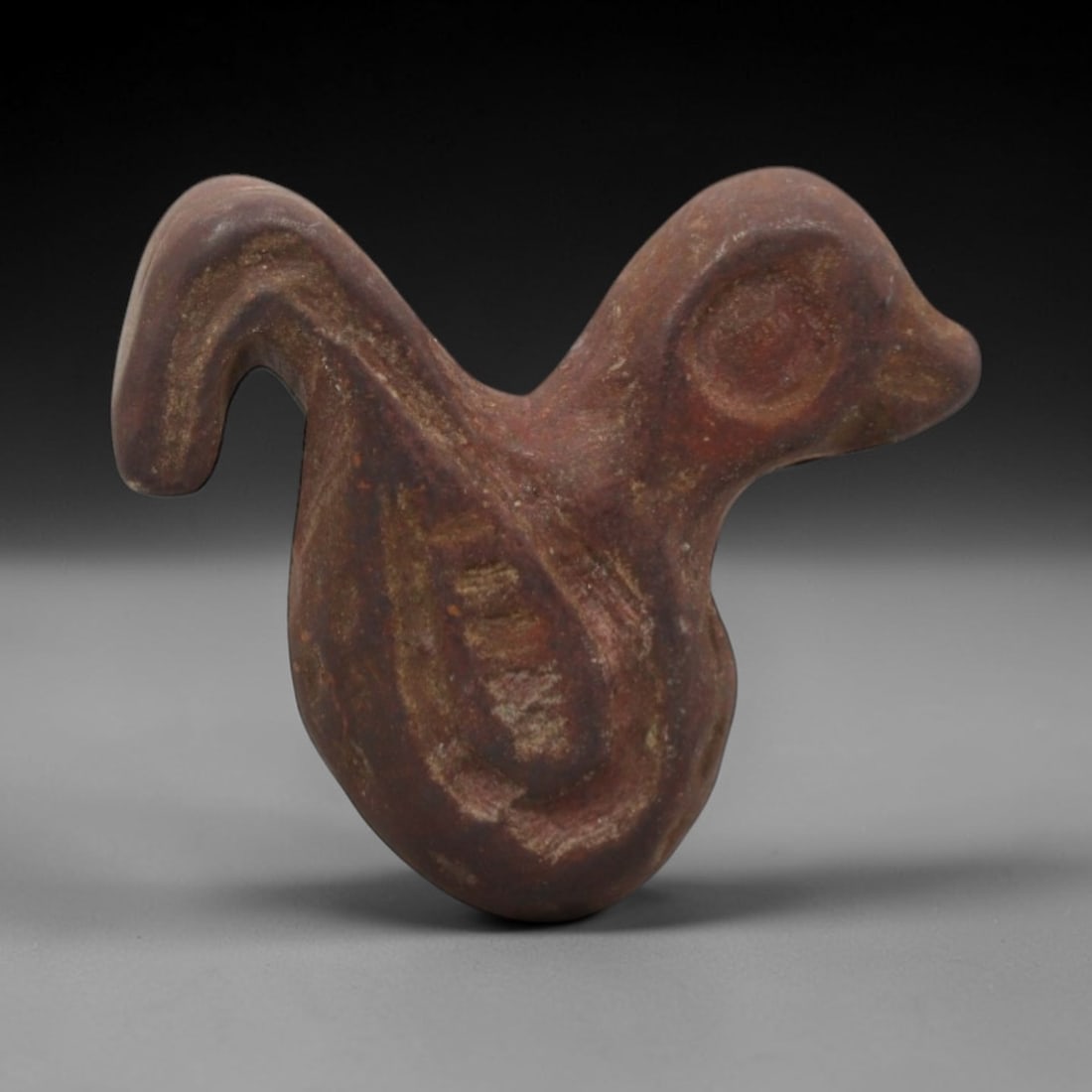 Ancient Near Eastern Stone Bird Amulet - Carved Figurine, 7ZV: This amulet is carved from a reddish-brown siltstone, with incised linear details defining its stylized avian form. The piece depicts a compact, stylized bird with a curved neck, rounded body, and inc