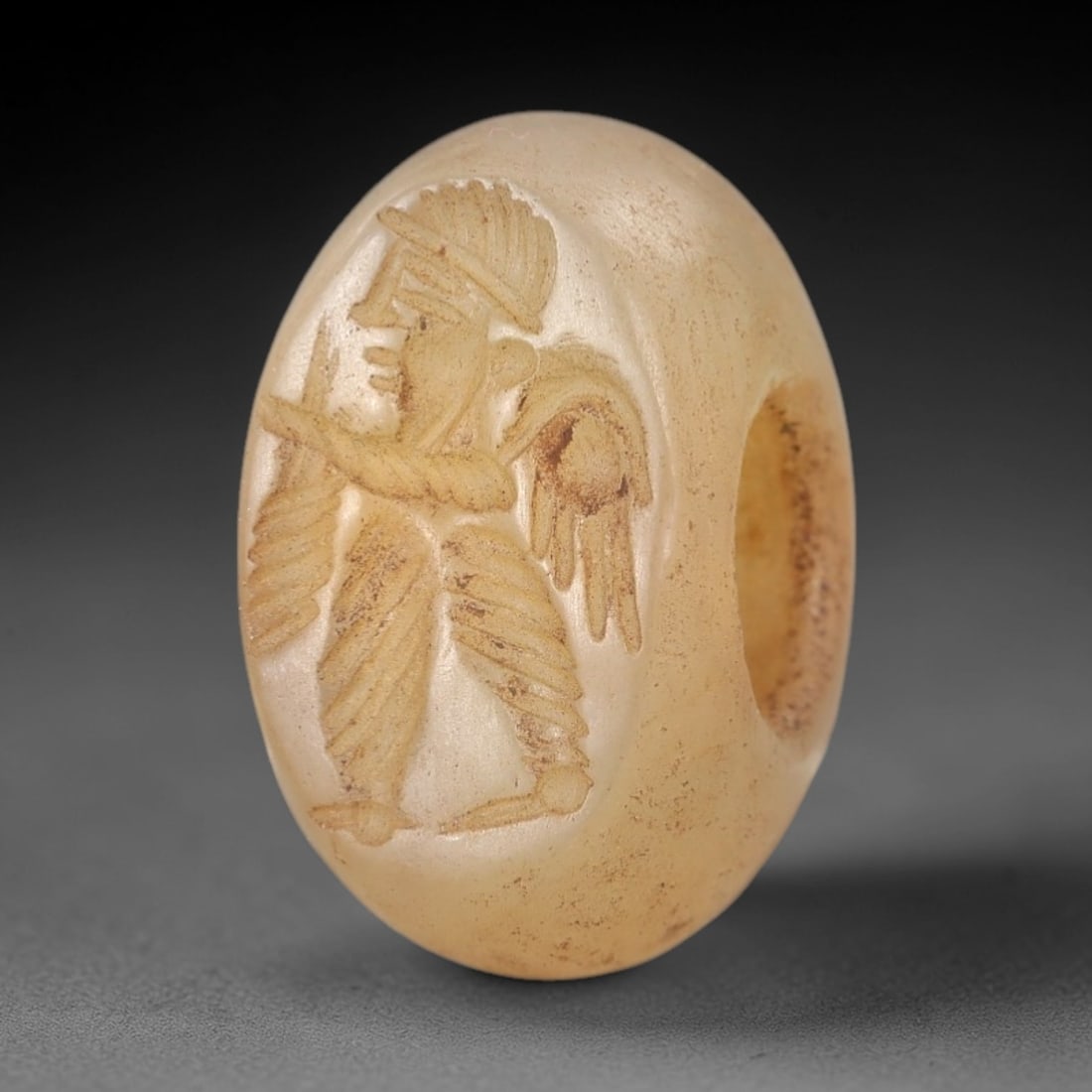 Ancient Near Eastern Chalcedony Stamp Seal - Winged Figure with Bow Motif, 98AM: This artifact is a hemispherical chalcedony stamp seal, executed in the classic glyptic tradition of the ancient Near East. The face features a striding winged figure clad in a pleated robe and creste