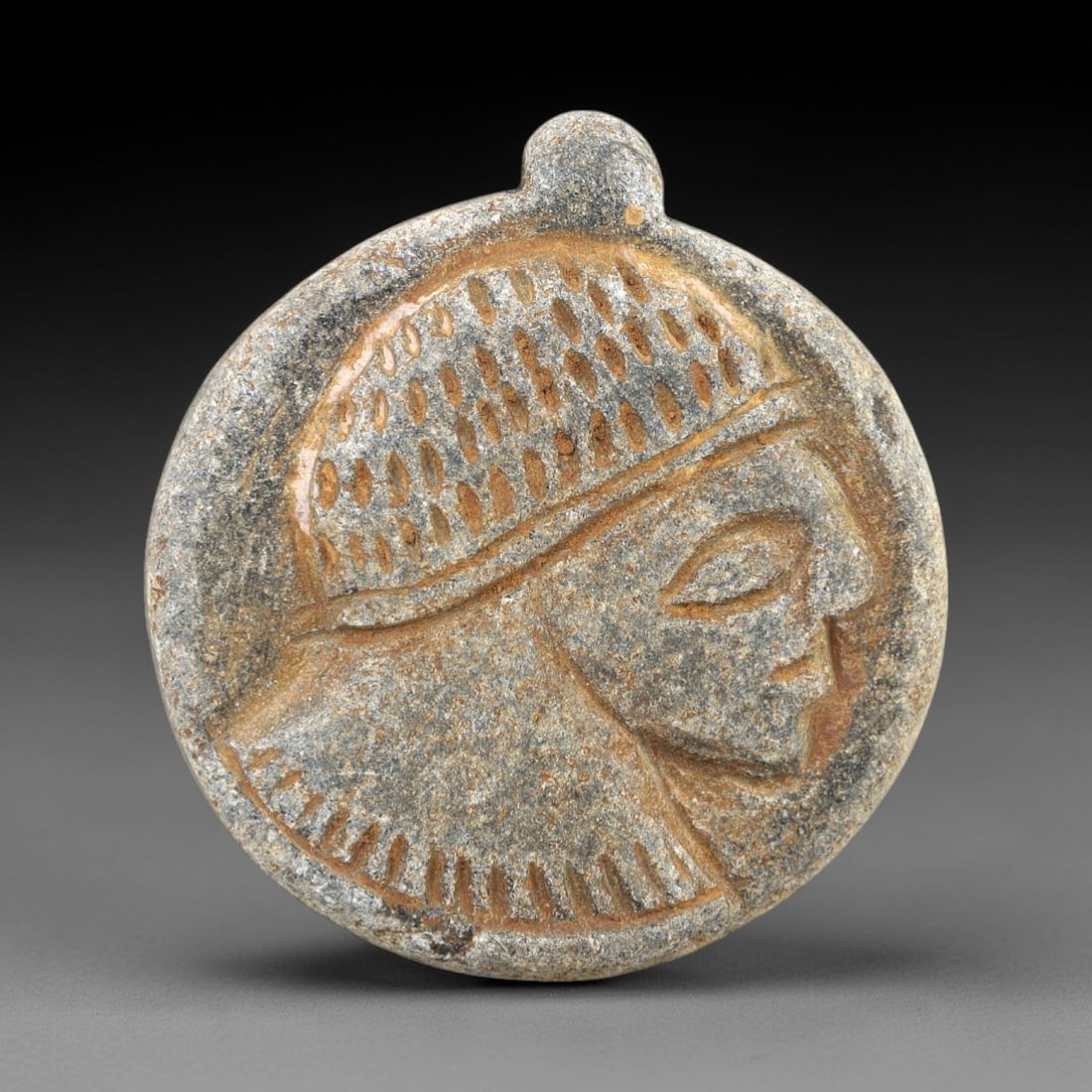 Ancient Stone Amulet - Near Eastern Portrait Pendant, 22AM: This circular pendant is carved from a weathered, mottled stone with a warm, earthy patina consistent with ancient Near Eastern burial contexts. The central relief depicts a stylized human profile, fe