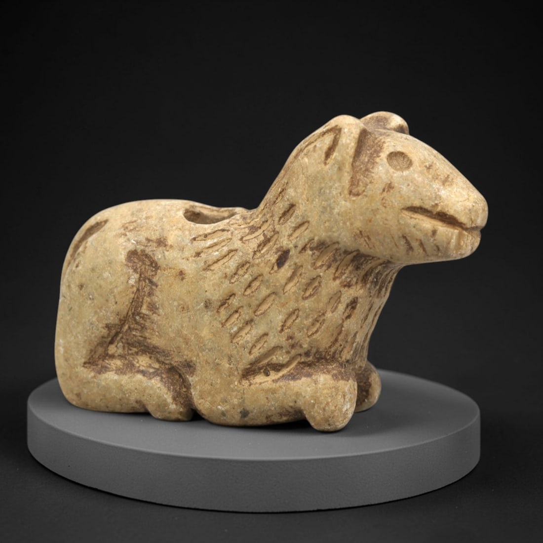 Ancient Stone Canine Figurine - Near Eastern Ritual Relics, 95ZP: Carved from warm tan stone, this reclining canine figurine features a stylized head with incised eyes, pointed ears, and a gently curved mouth. The neck and upper body display textured, linear incisio