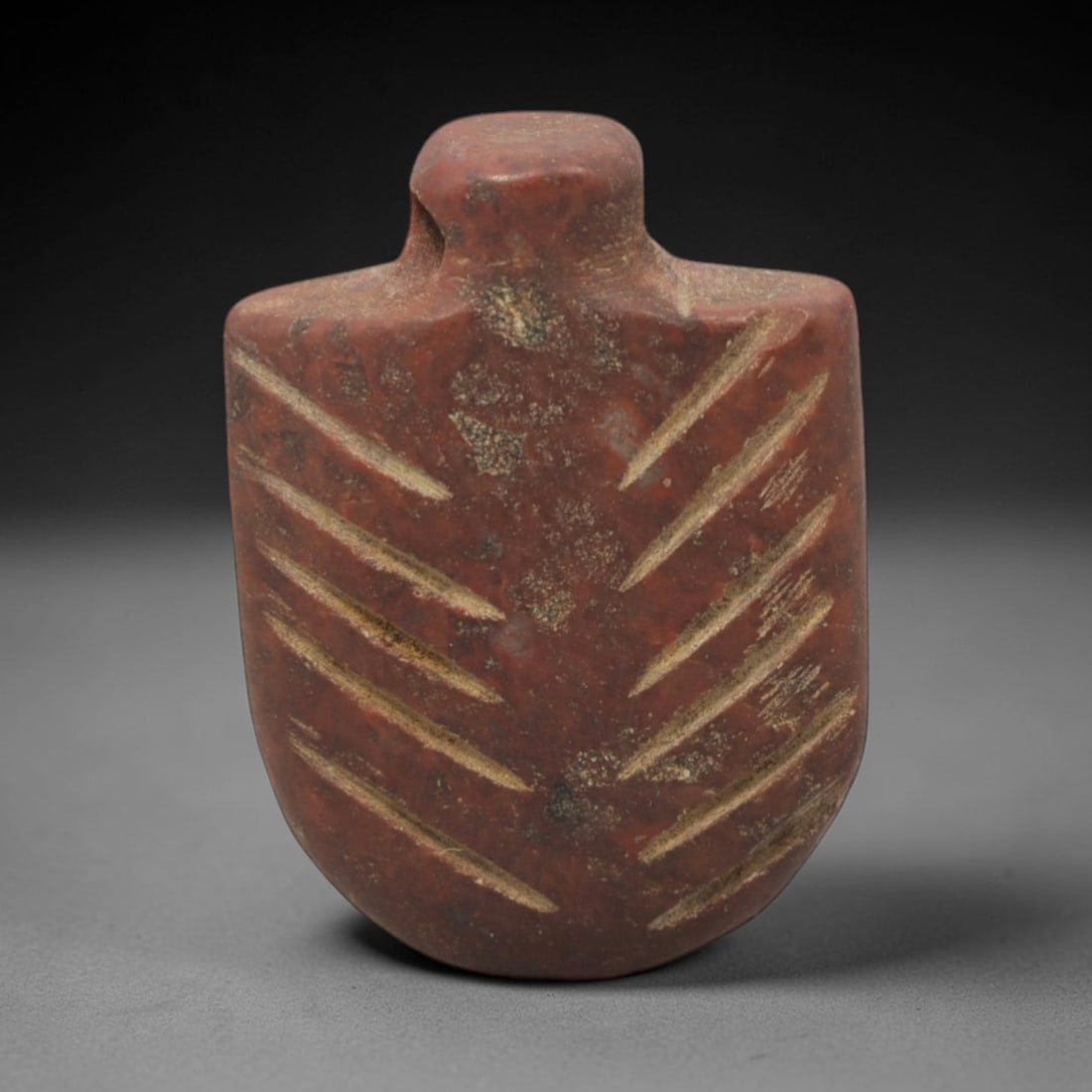 Ancient Stone Shield Amulet - Near Eastern Pendant, 15XR: Carved from a rich reddish stone, this amulet takes a shield-shaped form with a perforated top for suspension, its surface incised with symmetrical radiating linear patterns. The form and decoration a