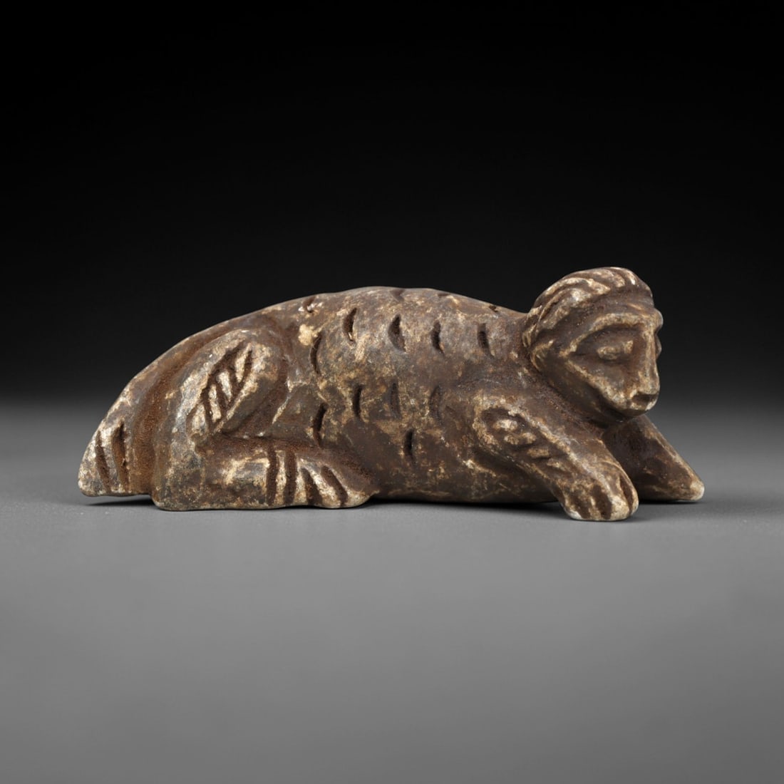 Ancient Schist Zoomorphic Amulet - Reclining Beast Figurine, 71ZP: This zoomorphic amulet is carved from schist, shaped into a reclining beast form with incised linear details defining the textured fur, stylized limbs, and facial features, including closed eyes and a