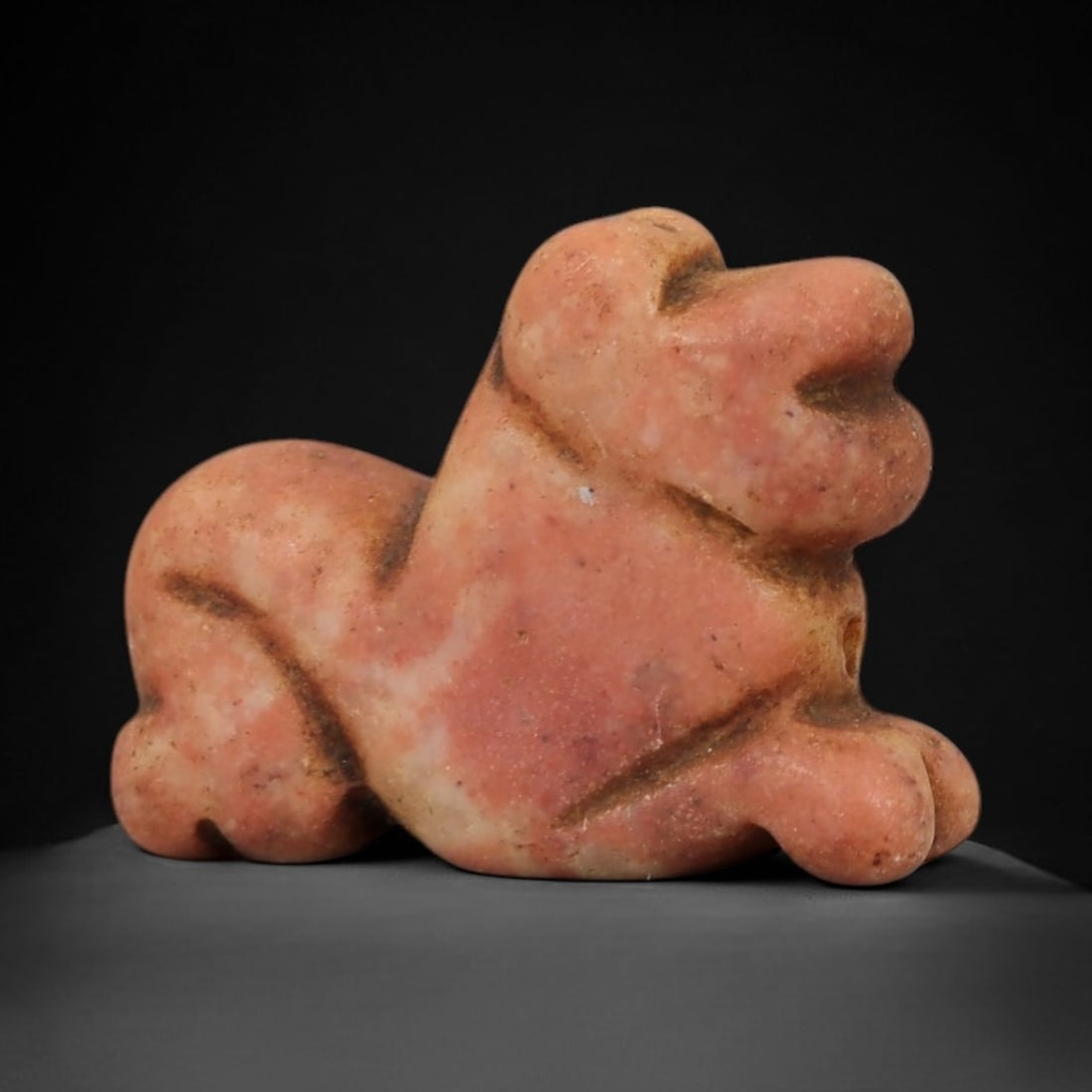Ancient Pink Stone Recumbent Feline - Near Eastern Amulet, 95AD: Carved from mottled pinkish-hued stone, this amulet depicts a recumbent feline in a compact, stylized form. The creature is rendered in a resting posture, with simplified facial features defined by in