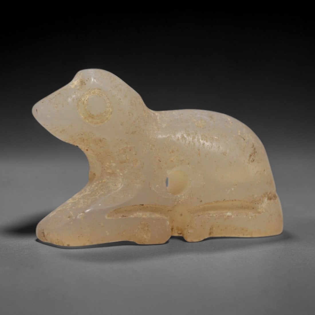 Ancient Chalcedony Recumbent Animal Amulet - Near Eastern Pendant, 80ZW: This amulet is carved from translucent pale chalcedony, depicting a stylized recumbent animal with simplified incised details defining the head, circular eye, and body contours. The creature is render