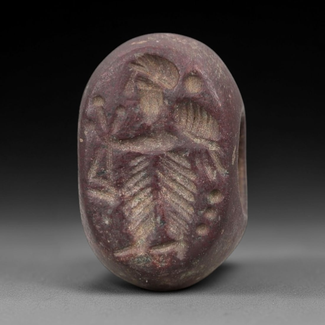 Ancient Red Stone Stamp Seal - Near Eastern Amulet, 36AN: Crafted from dense, reddish-purple stone, this hemispherical stamp seal displays incised iconography of a standing male figure in profile, adorned with a headdress and full beard, clad in a layered, f