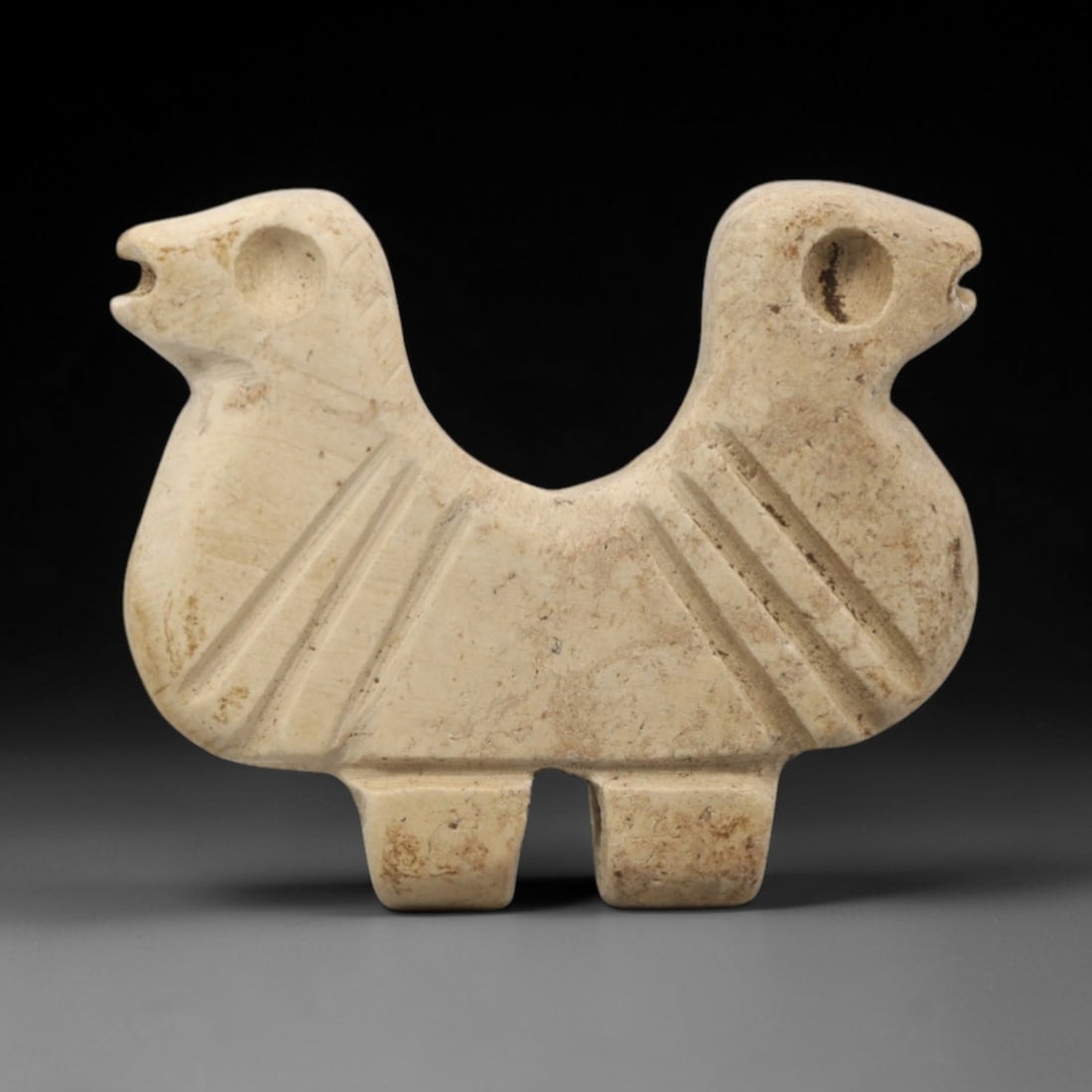 Ancient Near Eastern Stone Double Bird Amulet - Carved Figurine, 90ZW: This amulet is carved from a pale, creamy limestone, with incised linear details defining its symmetrical double-bird form. The piece features two stylized bird heads facing outward, each with a circu