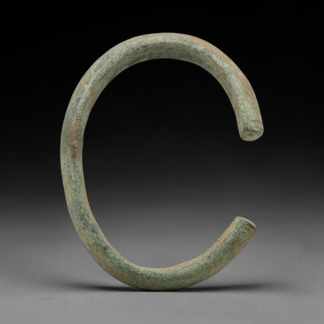 A Roman Bronze Bracelet, 1st-3rd Century CE, 9FV: Cast from bronze with a thick, rounded section, the bracelet is formed as an open C-shape with flat, slightly tapered terminals, each marked by a shallow incised line. The surface is covered in a unif