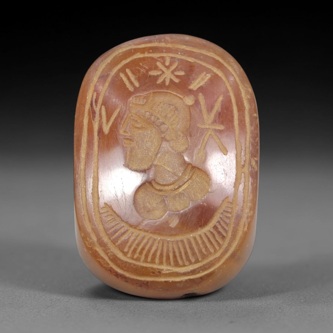Ancient Carnelian Intaglio - Near Eastern Figural Seal, 65ZP: This oval intaglio is carved from polished carnelian, featuring a bearded male bust in profile, adorned with a head covering and necklet, surrounded by symbolic linear and star-like motifs within a bo