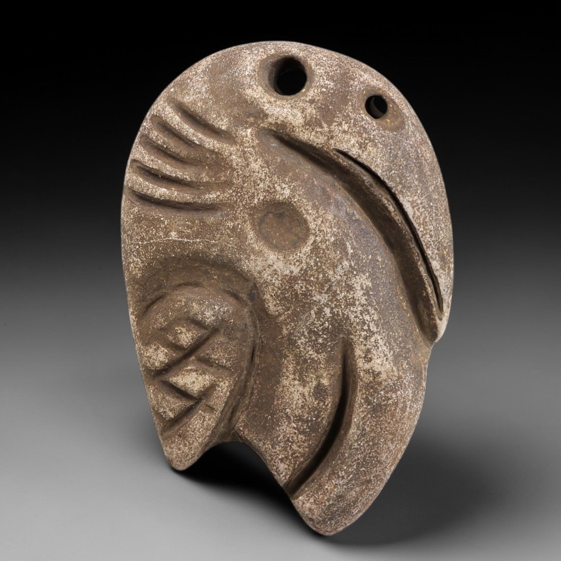 Ancient Stone Bird Amulet - Near Eastern Zoomorphic Pendant, 93FX: Carved from a weathered granular stone, this amulet depicts a stylized bird head in profile, with incised details defining the beak, feathered crest, and geometric body motifs. Two perforations at the