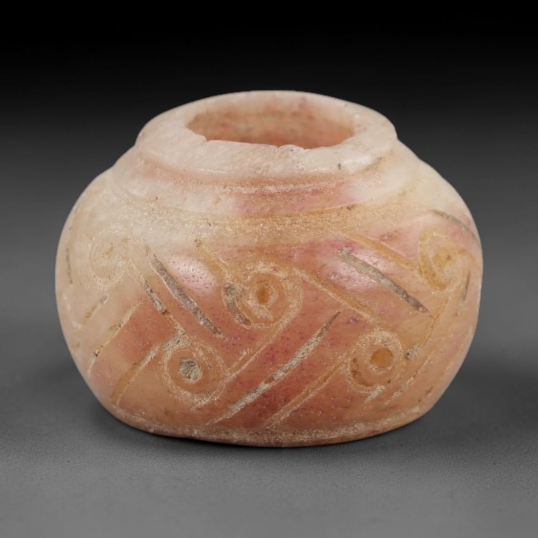 Ancient Stone Spindle Whorl - Near Eastern Spiral Motif, 80XR: This ancient Near Eastern spindle whorl is carved from stone, featuring incised spiral and linear geometric patterns surrounding the central perforation. The compact, rounded form is a traditional tex